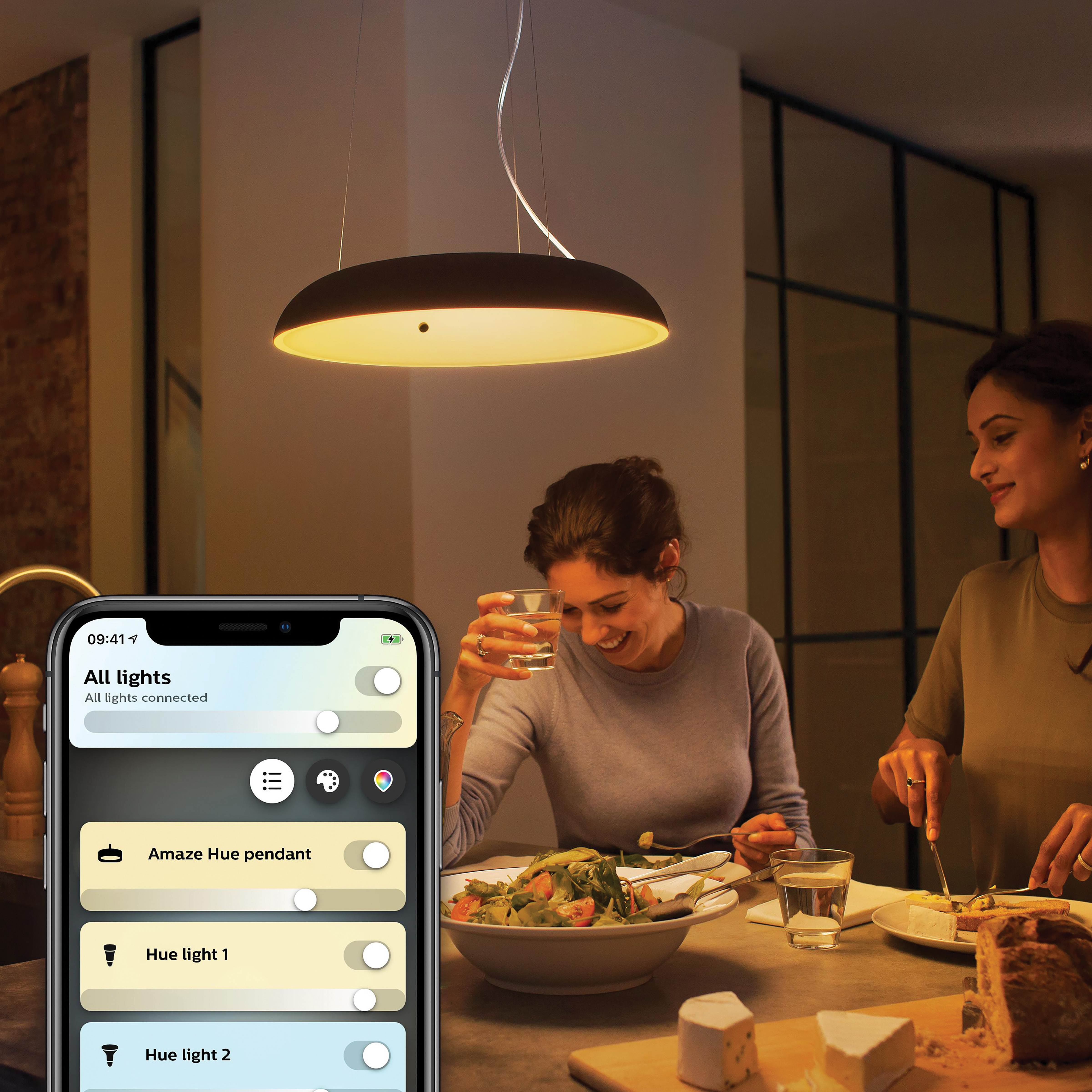 Deux personnes sont à table, l'une tient un verre d'eau. Un smartphone affiche une application de maison intelligente contrôlant les lumières.
