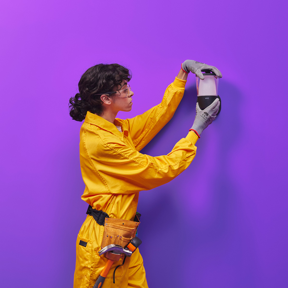 Une personne en combinaison jaune installe un luminaire noir sur un mur violet.