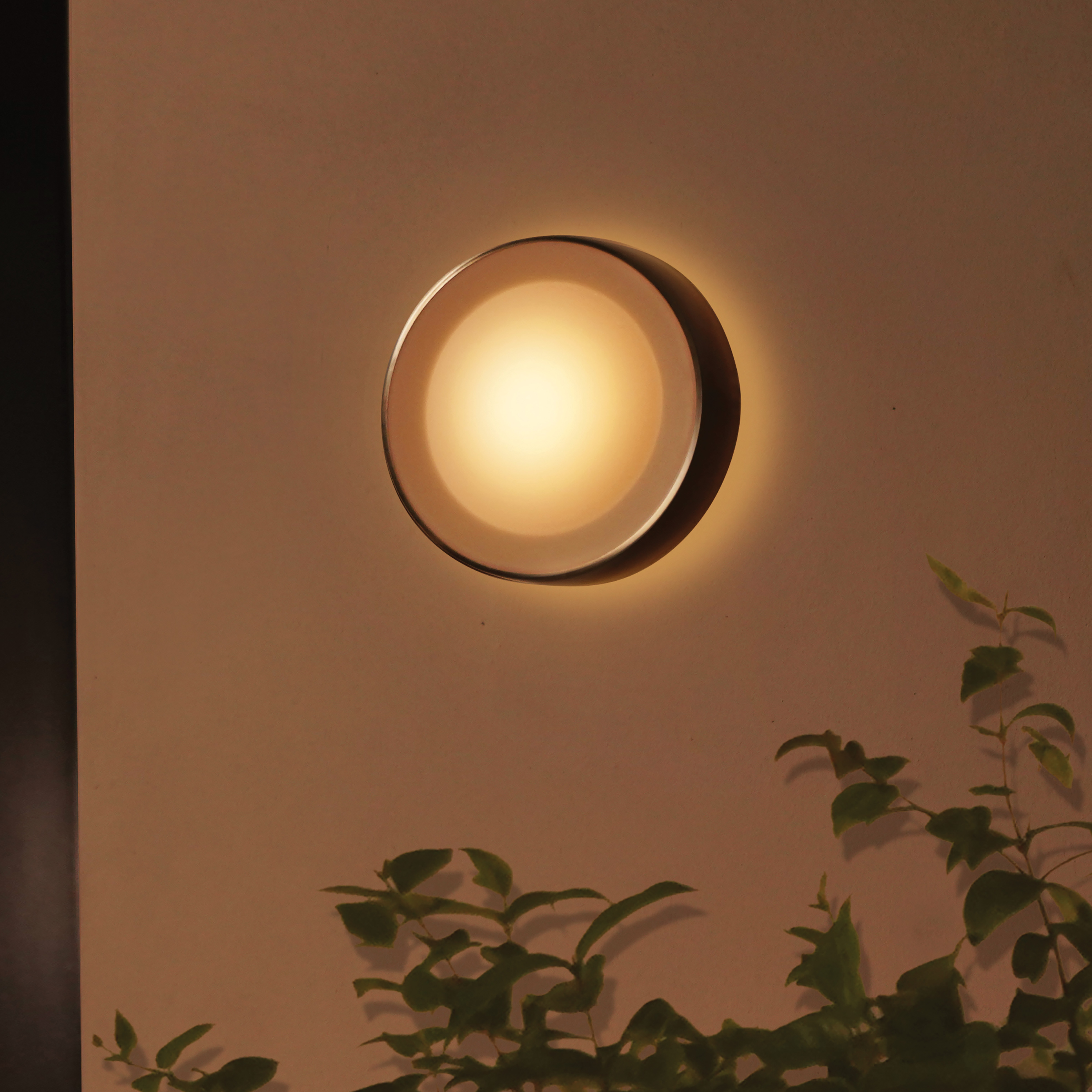Een cirkelvormige wandlamp wordt getoond. Hij is verlicht, met een warme gloed. Groene bladeren staan onder in de afbeelding.