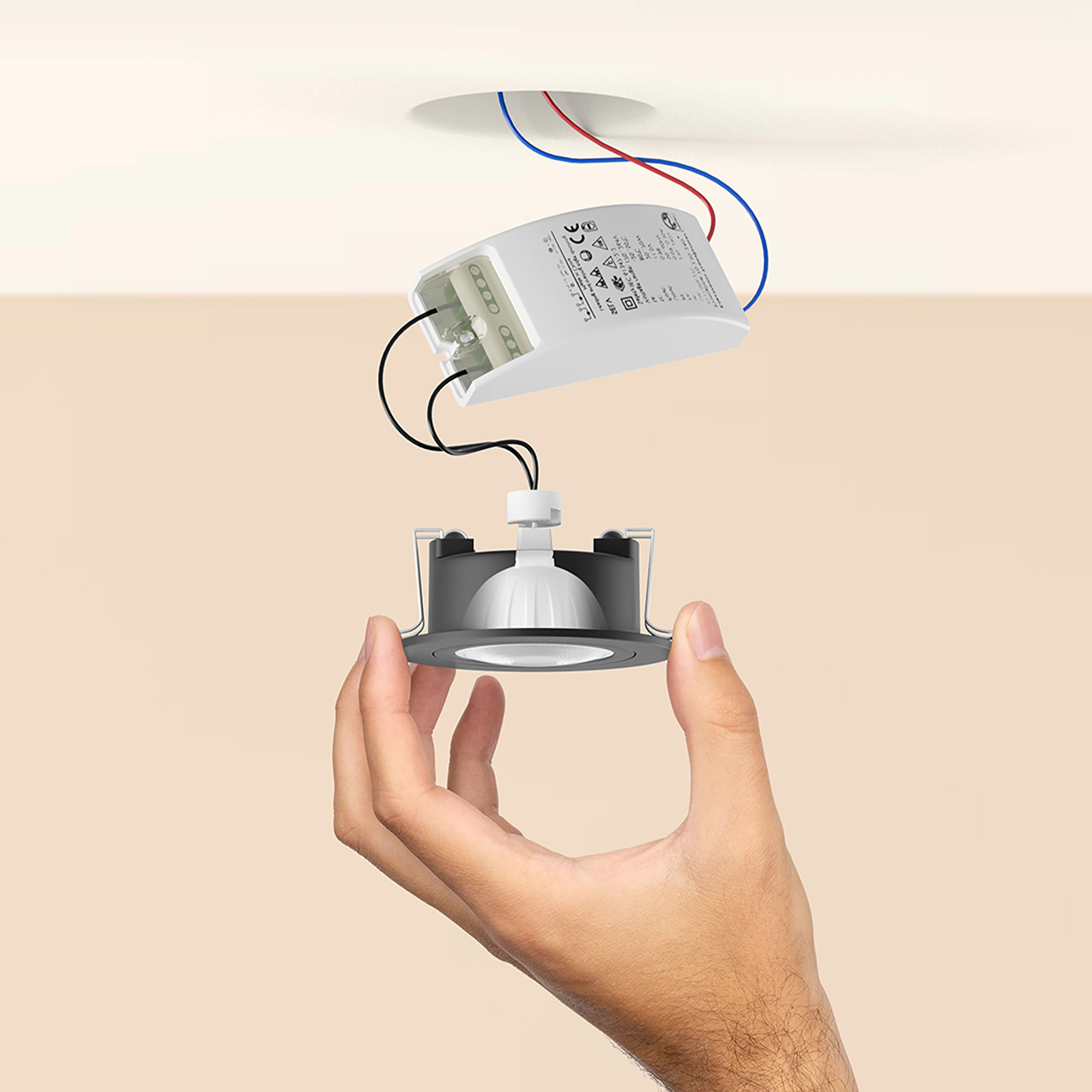 Ręka trzymająca oprawę wpuszczaną. Przewody i białe pudełko znajdują się nad oprawą. Tło jest beżowe.