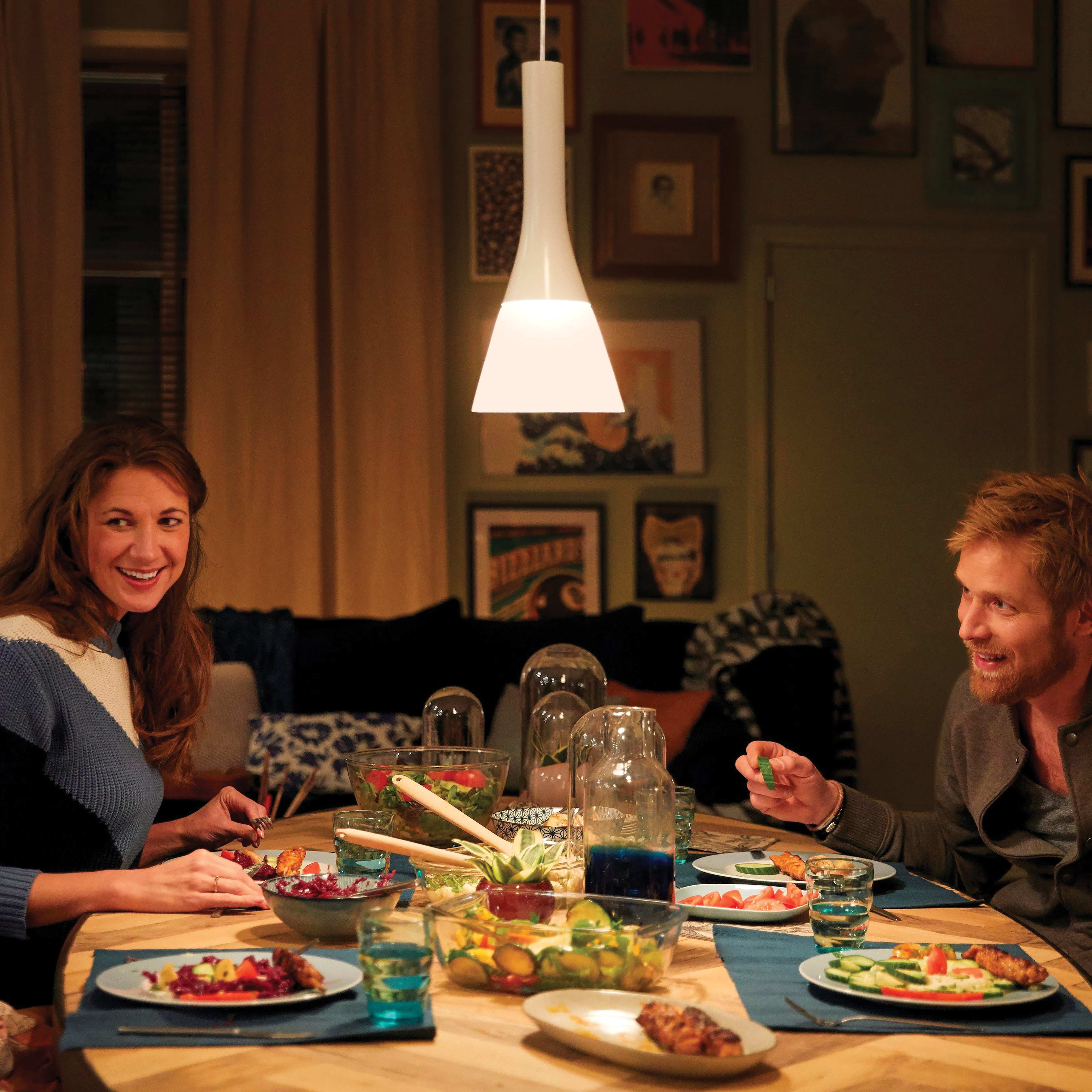 Ludzie przy stole z jedzeniem i napojami. Wisząca lampa oświetla scenę.