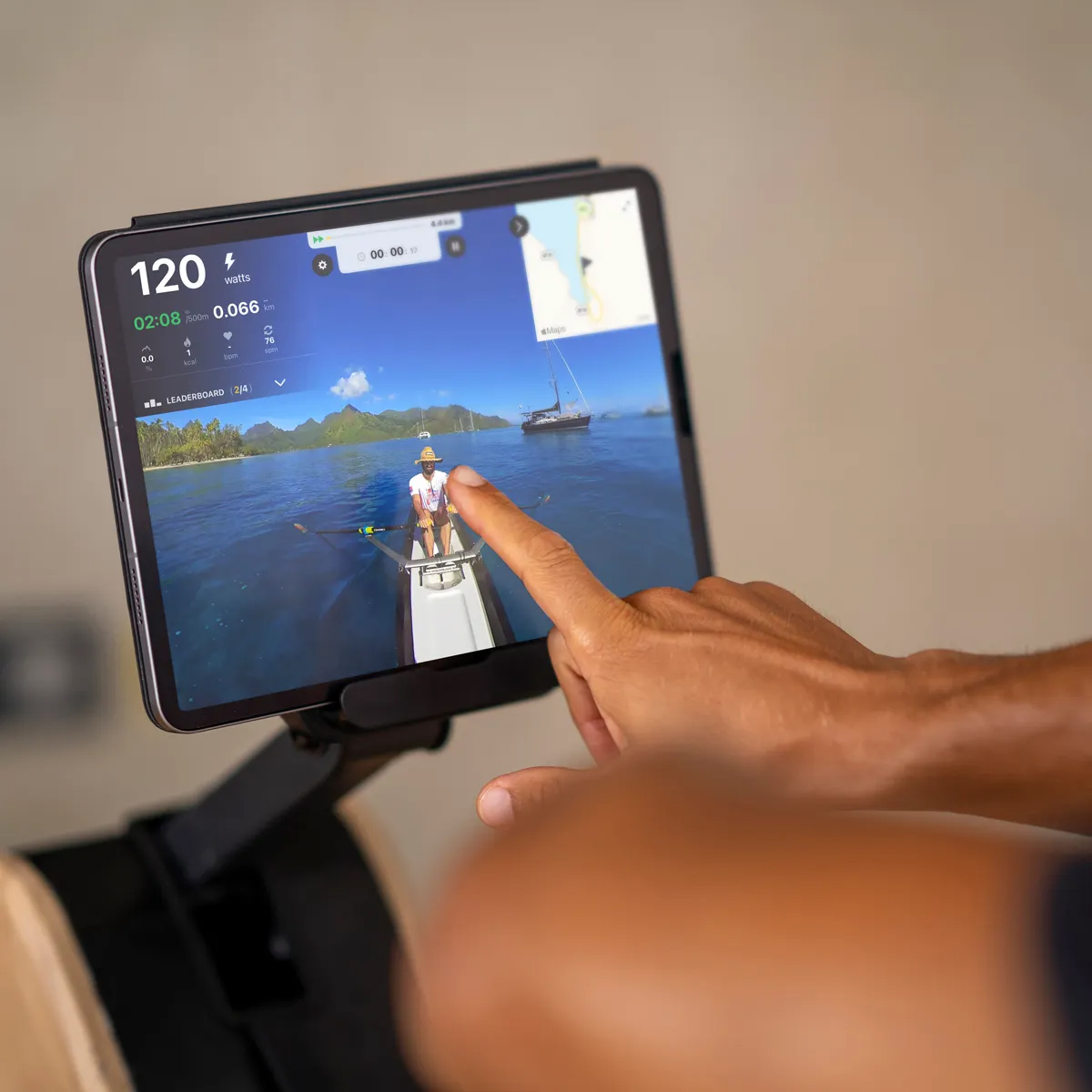 Die Hand einer Person berührt einen Tablet-Bildschirm, der eine Ruderszene zeigt. Das Wasser und Boote sind sichtbar.