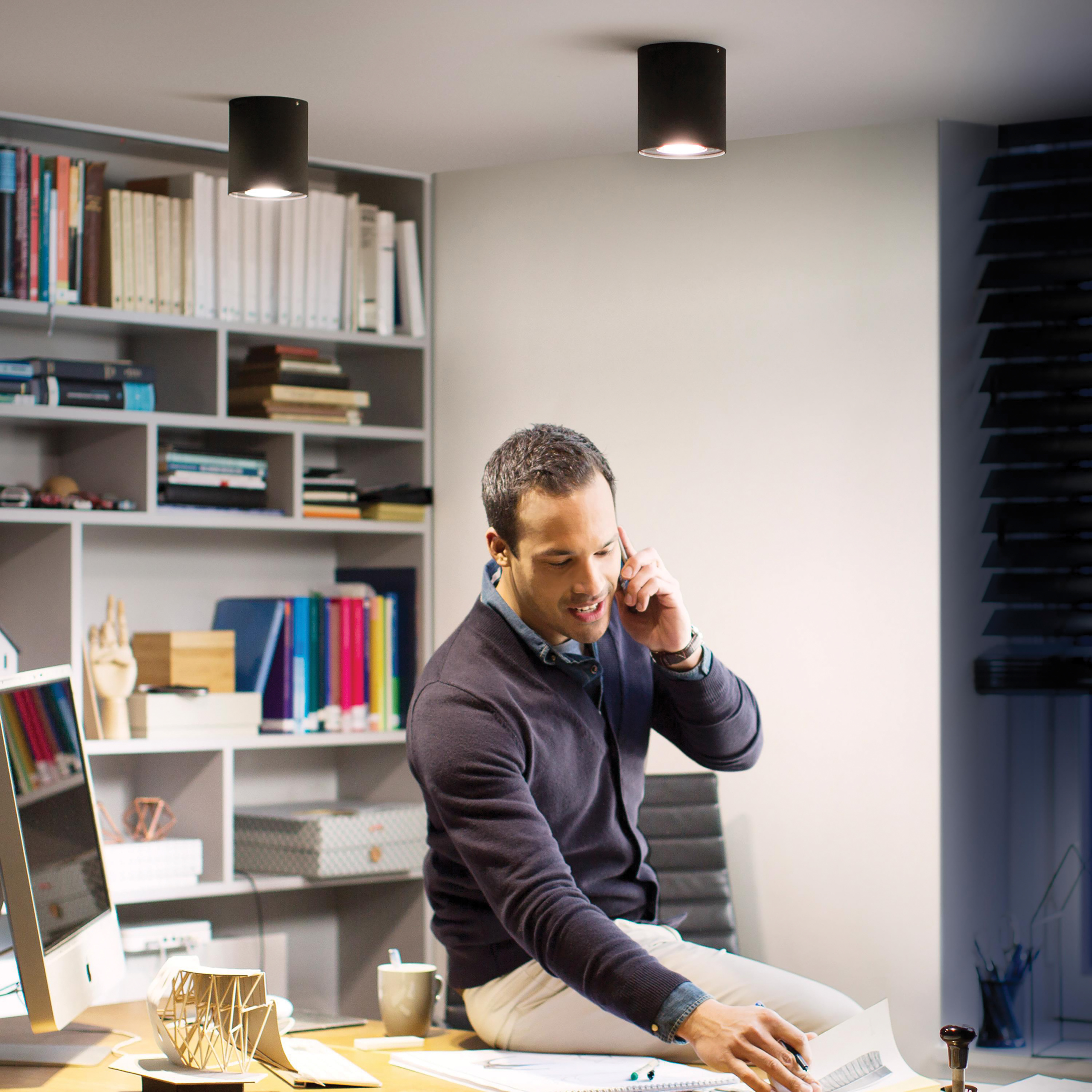 Mężczyzna przez telefon w biurze z dwoma lampami sufitowymi, półkami i oknem.