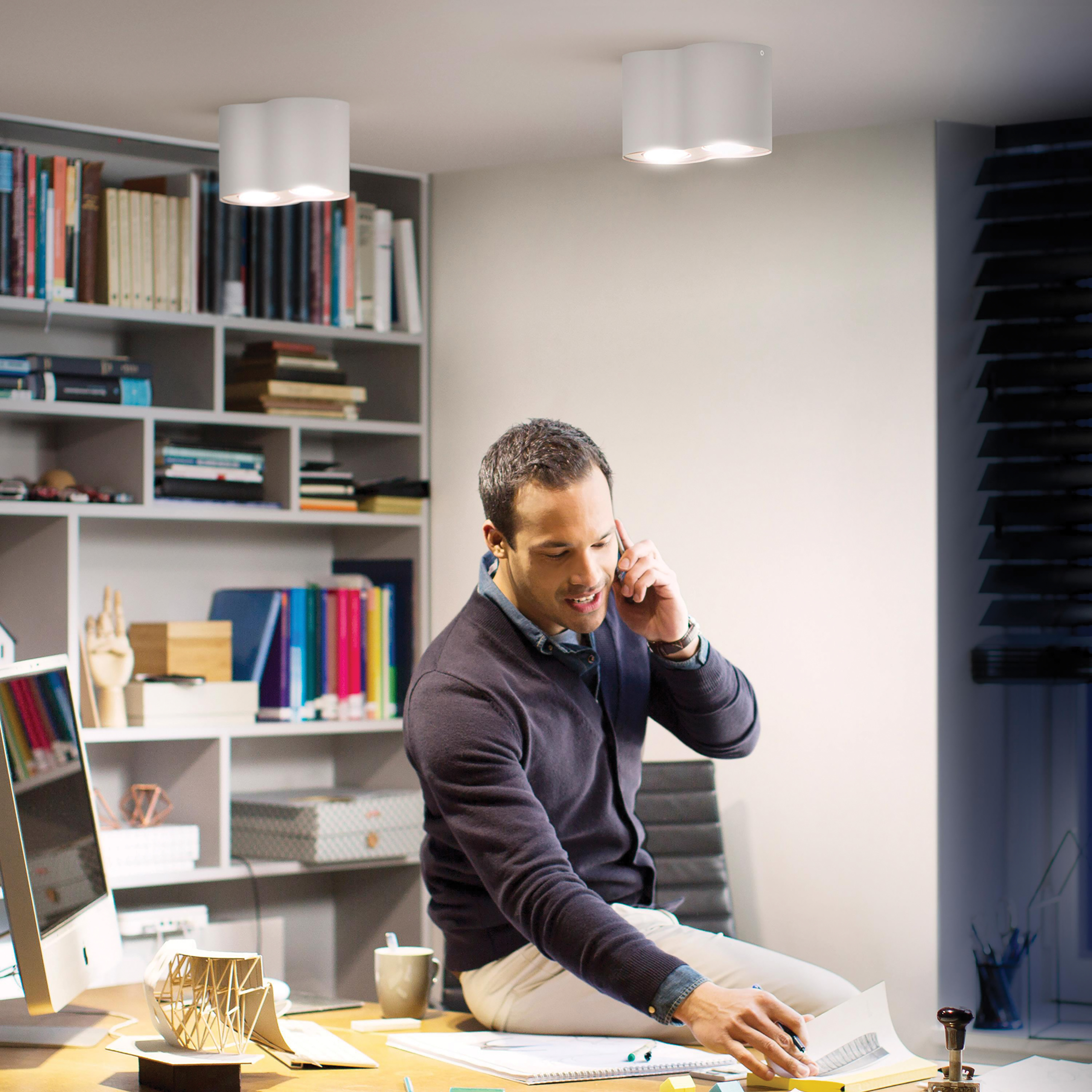 Mężczyzna w biurze rozmawia przez telefon, siedząc na biurku, z dwoma światłami nad nim.