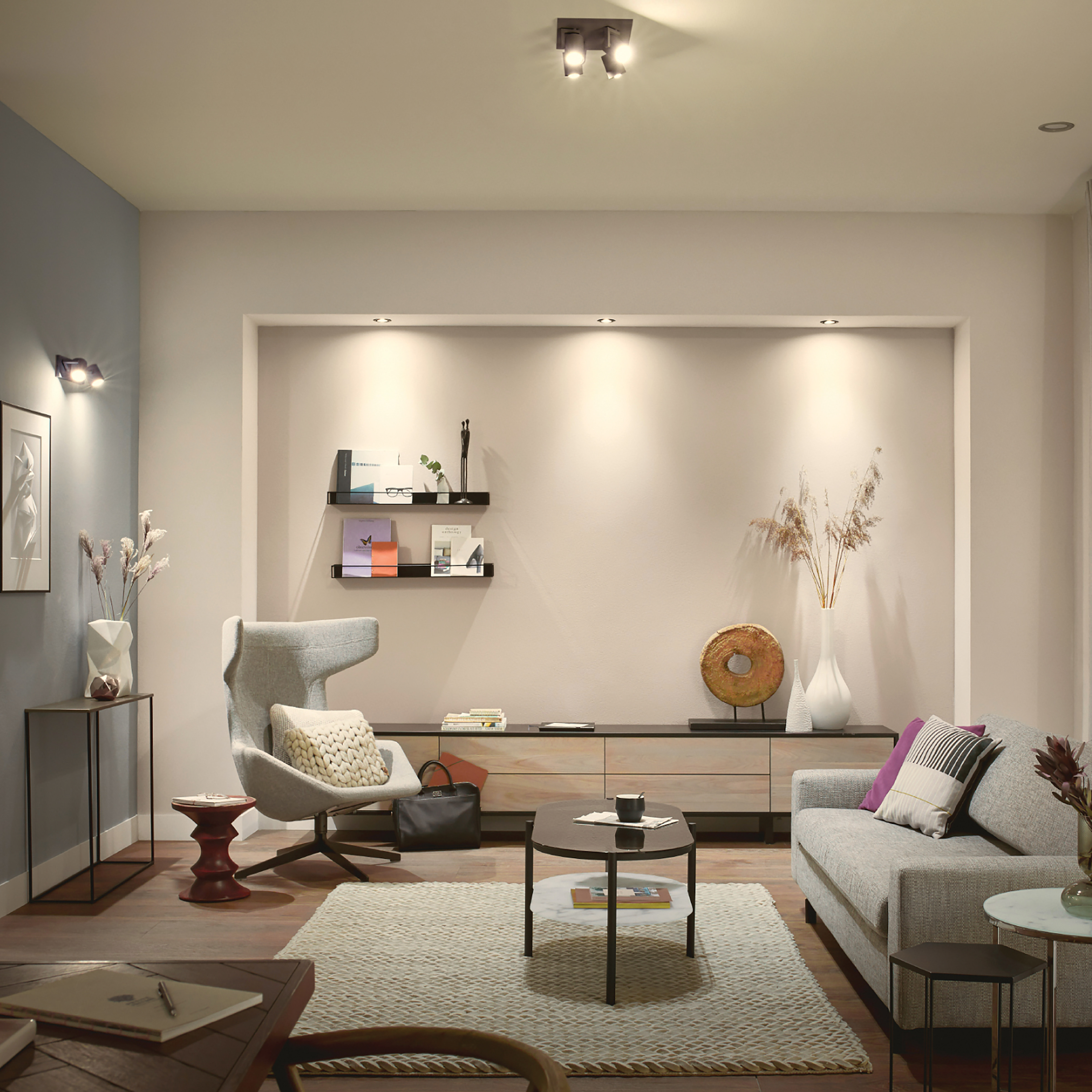 Salon z szarą sofą, dziełami sztuki, książkami, stolikiem bocznym i dwoma lampami sufitowymi.