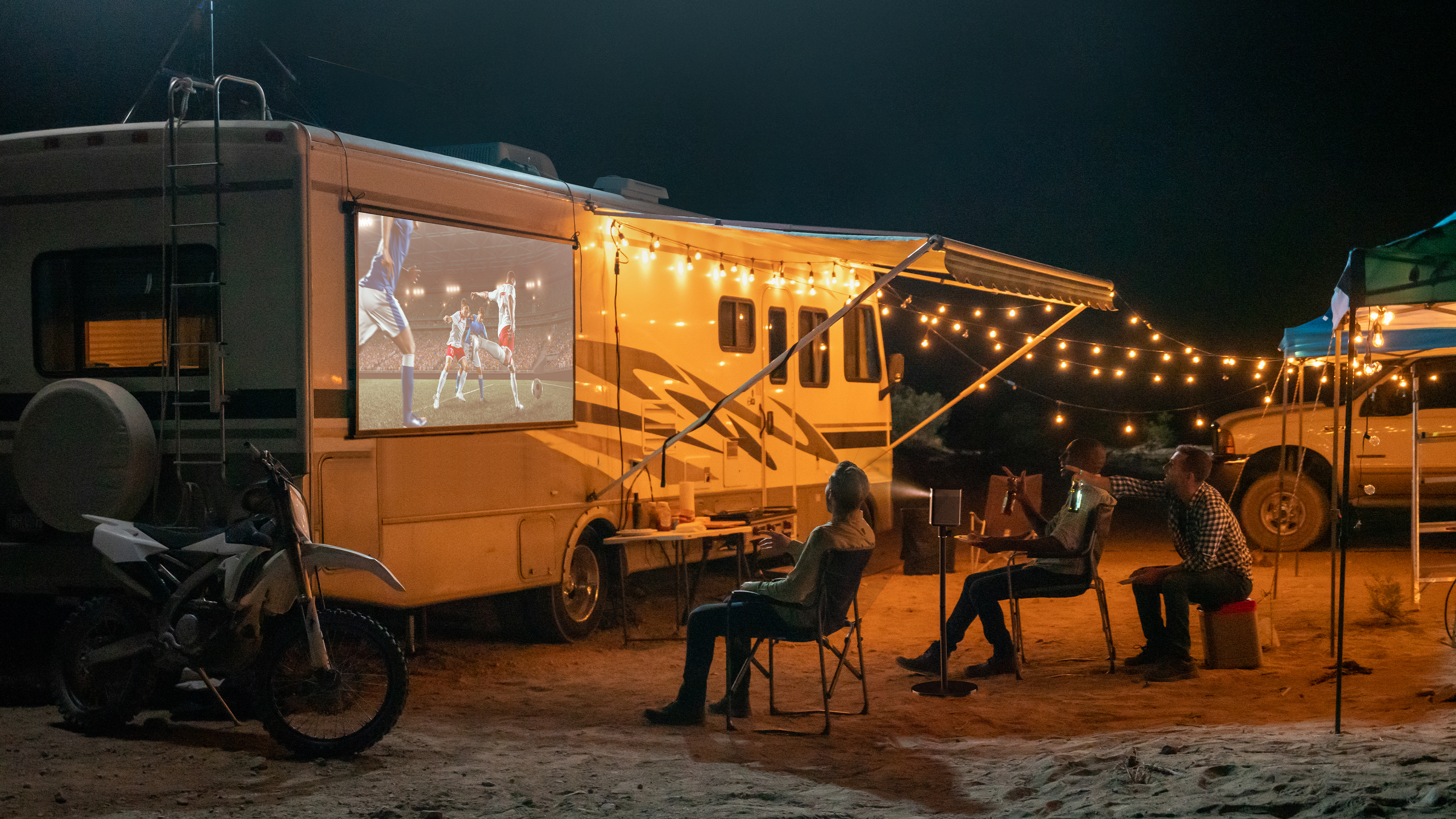 Grupo de personas viendo una película proyectada en una pantalla fuera de una autocaravana por la noche.