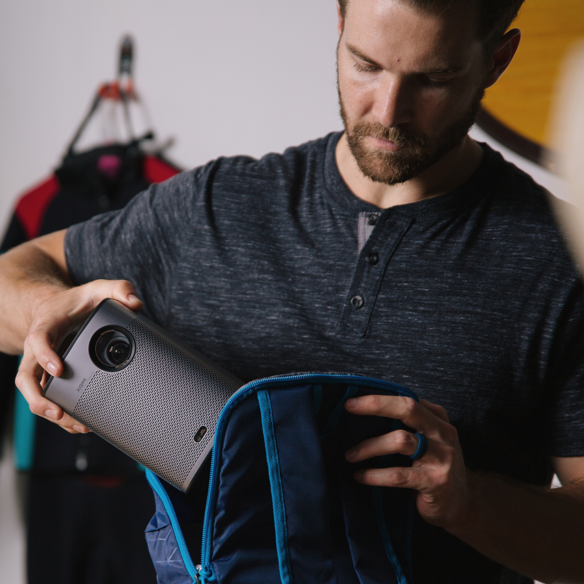 Hombre metiendo un proyector gris en una mochila azul. Fondo con ropa en una percha.