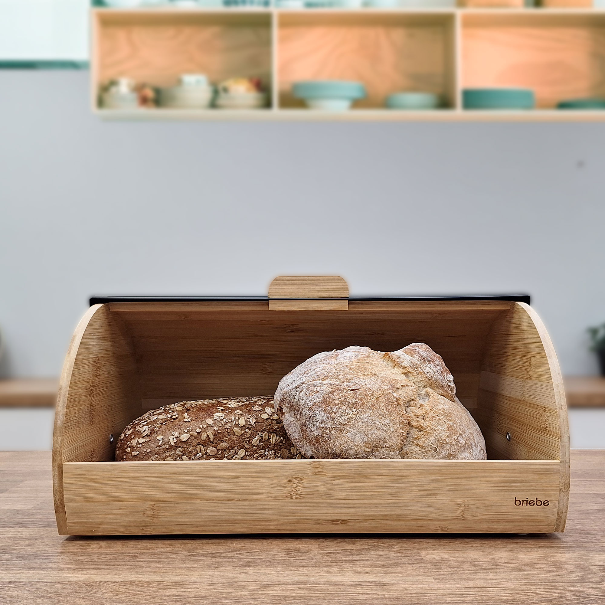 Caja de pan de madera con dos hogazas dentro. La caja está sobre una mesa de madera. Estanterías al fondo.