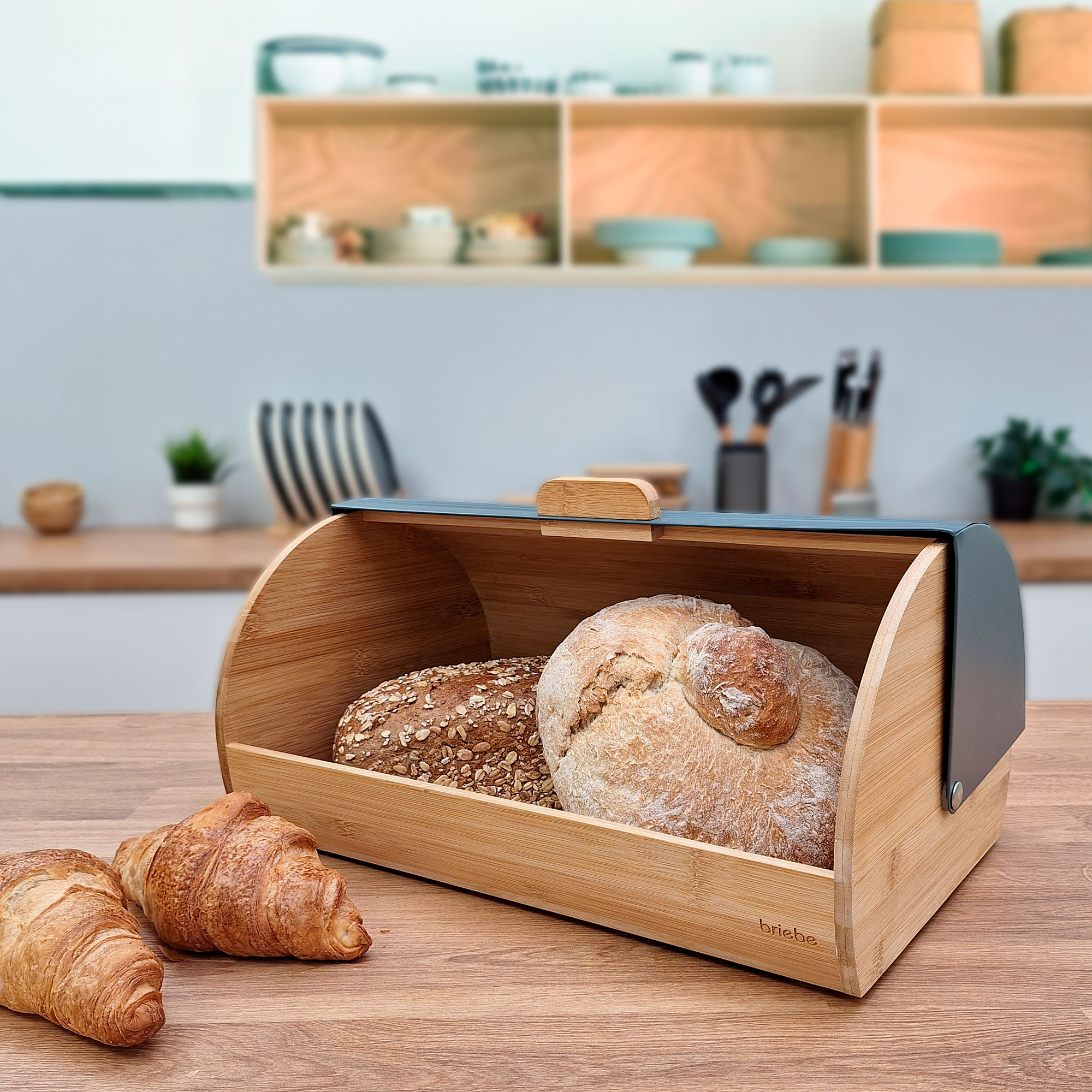 Una panera de madera con dos hogazas de pan y cruasanes dentro, colocada sobre una mesa de madera.