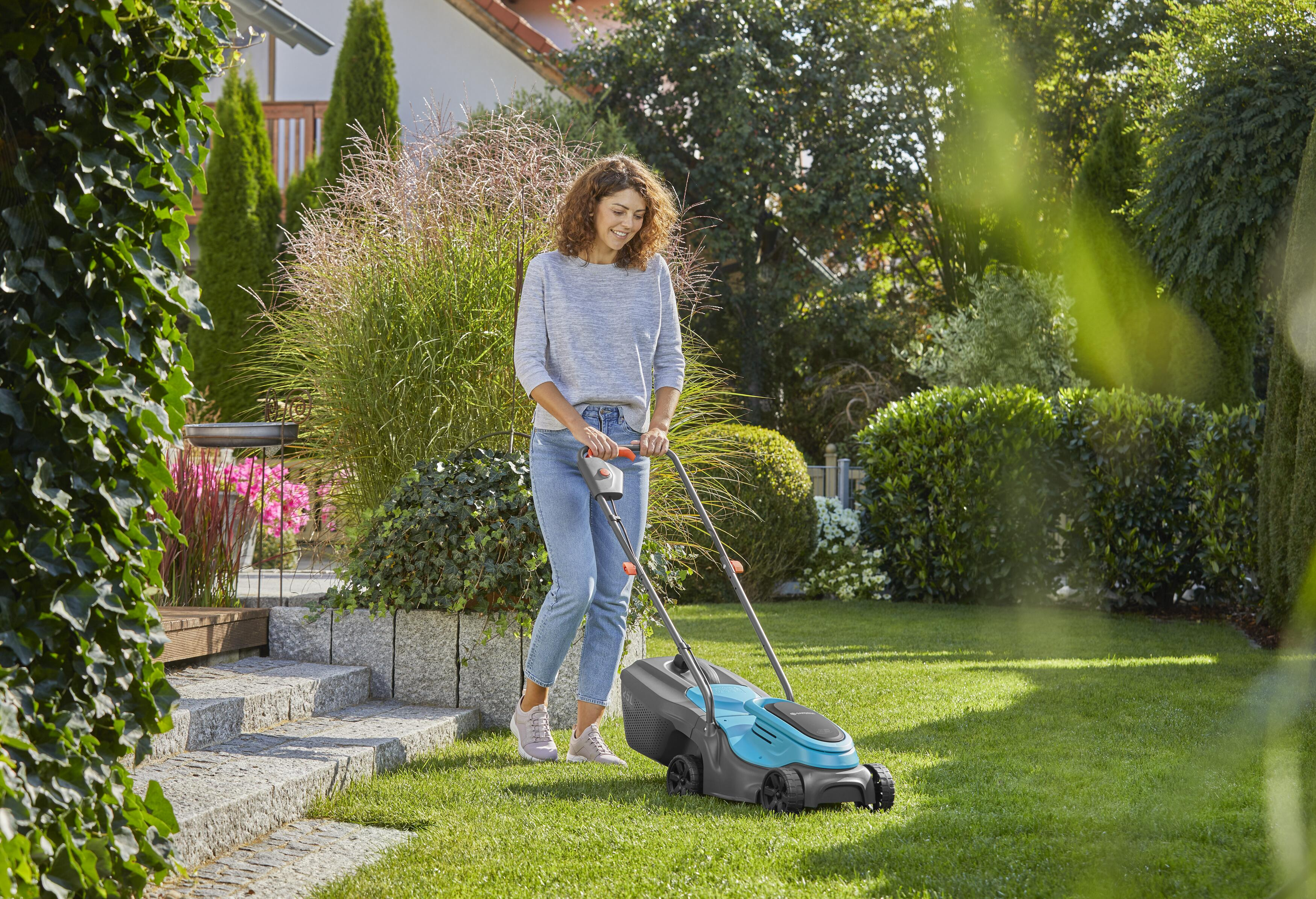Eine Frau mäht an einem sonnigen Tag in einem Garten einen grünen Rasen mit einem blau-schwarzen Rasenmäher.
