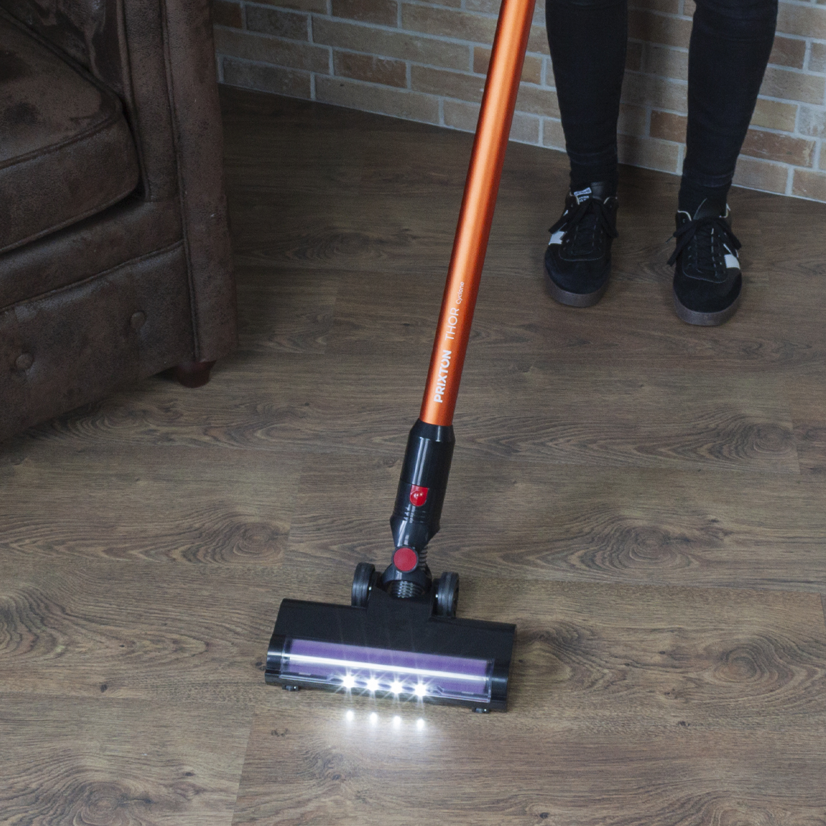 Osoba odkurza drewnianą podłogę pomarańczowo-czarnym odkurzaczem bezprzewodowym. Podłoga jest czysta.