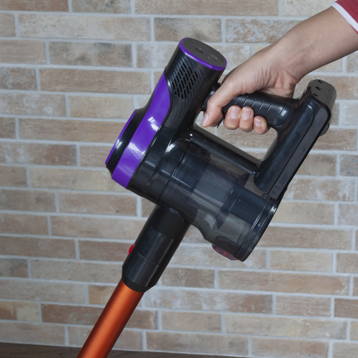 Osoba trzymająca czarno-fioletowy bezprzewodowy odkurzacz, blisko ceglanej ściany.