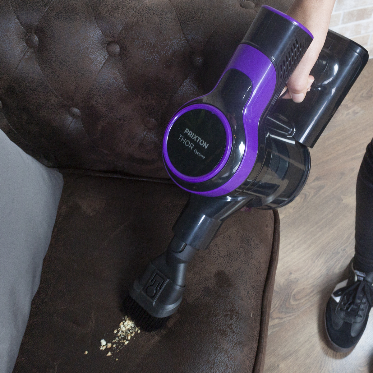 Osoba odkurzająca brązową sofę czarno-fioletowym odkurzaczem ręcznym.