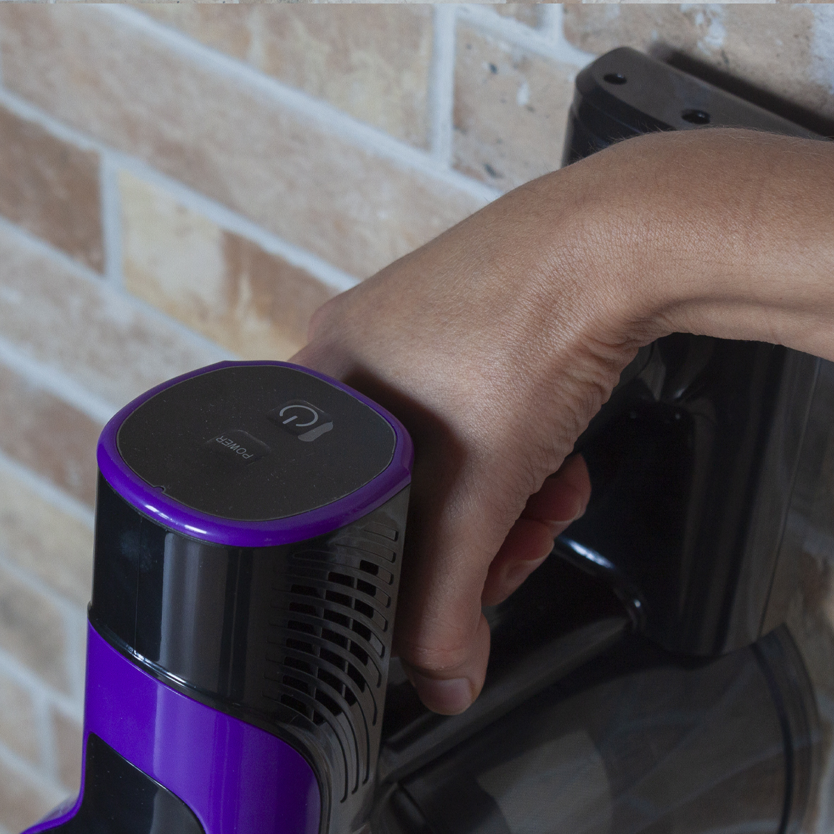 Ręka trzymająca fioletowo-czarny odkurzacz w pobliżu ceglanej ściany.