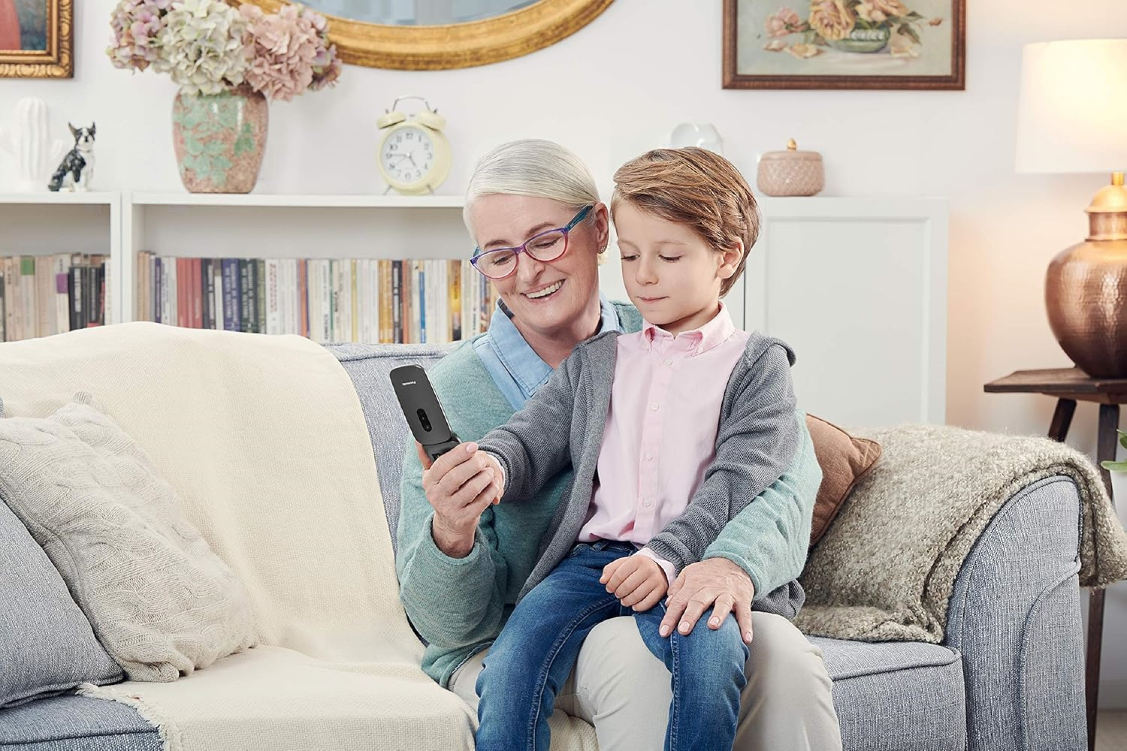 Großmutter und Kind auf einem Sofa, schauen gemeinsam auf ein Klapphandy, lächeln.