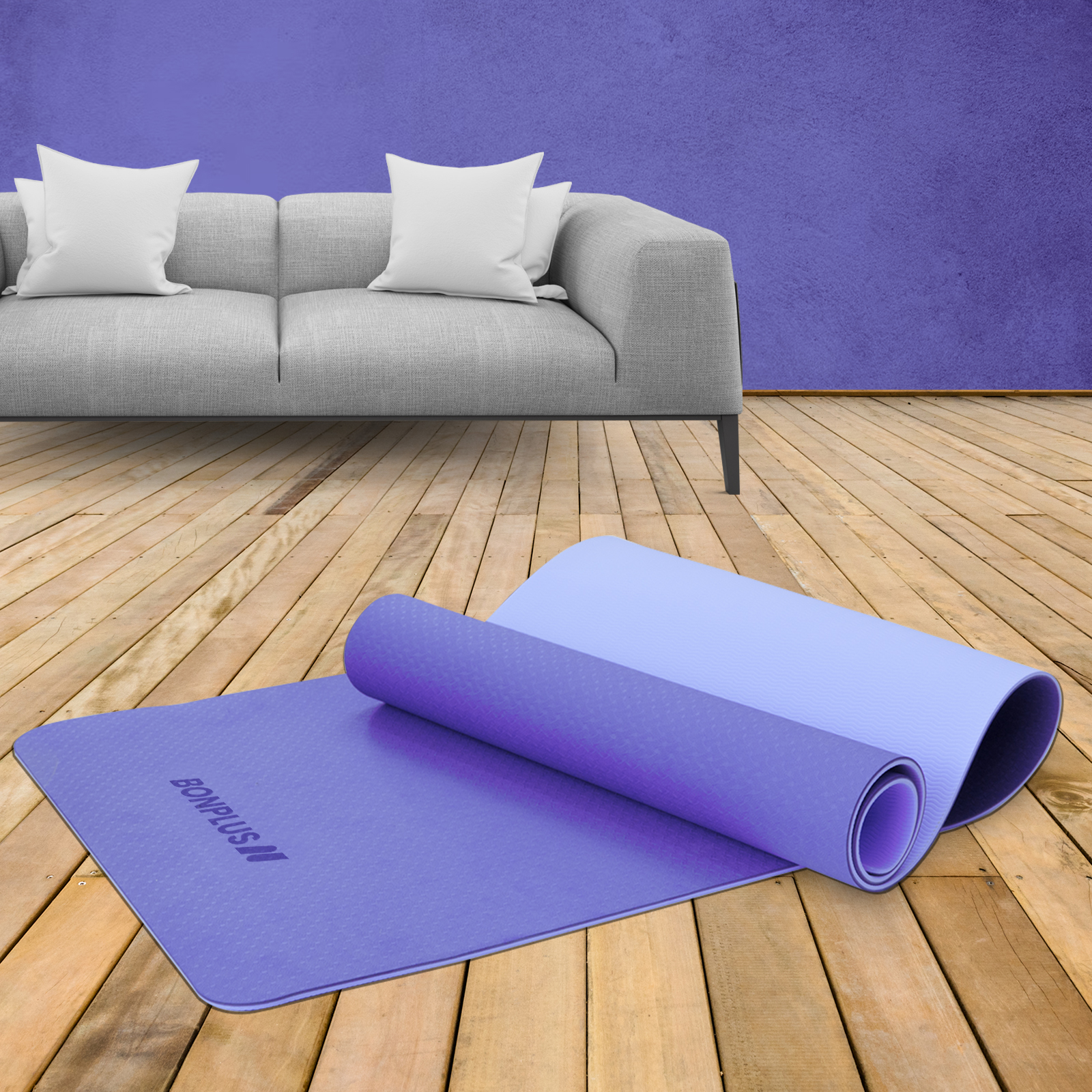 Una esterilla de yoga morada enrollada está en un suelo de madera junto a una extendida. Sofá gris al fondo.