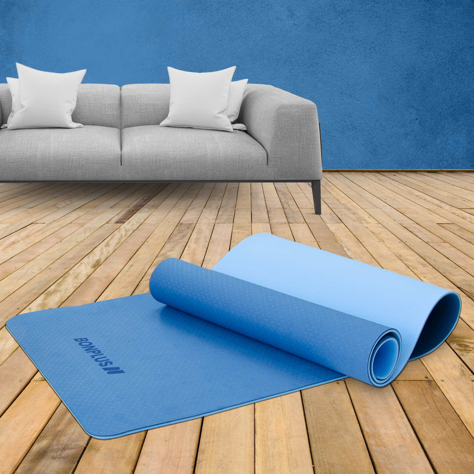 Esterilla de yoga azul enrollada y abierta en suelo de madera. Sofá gris y pared azul de fondo.