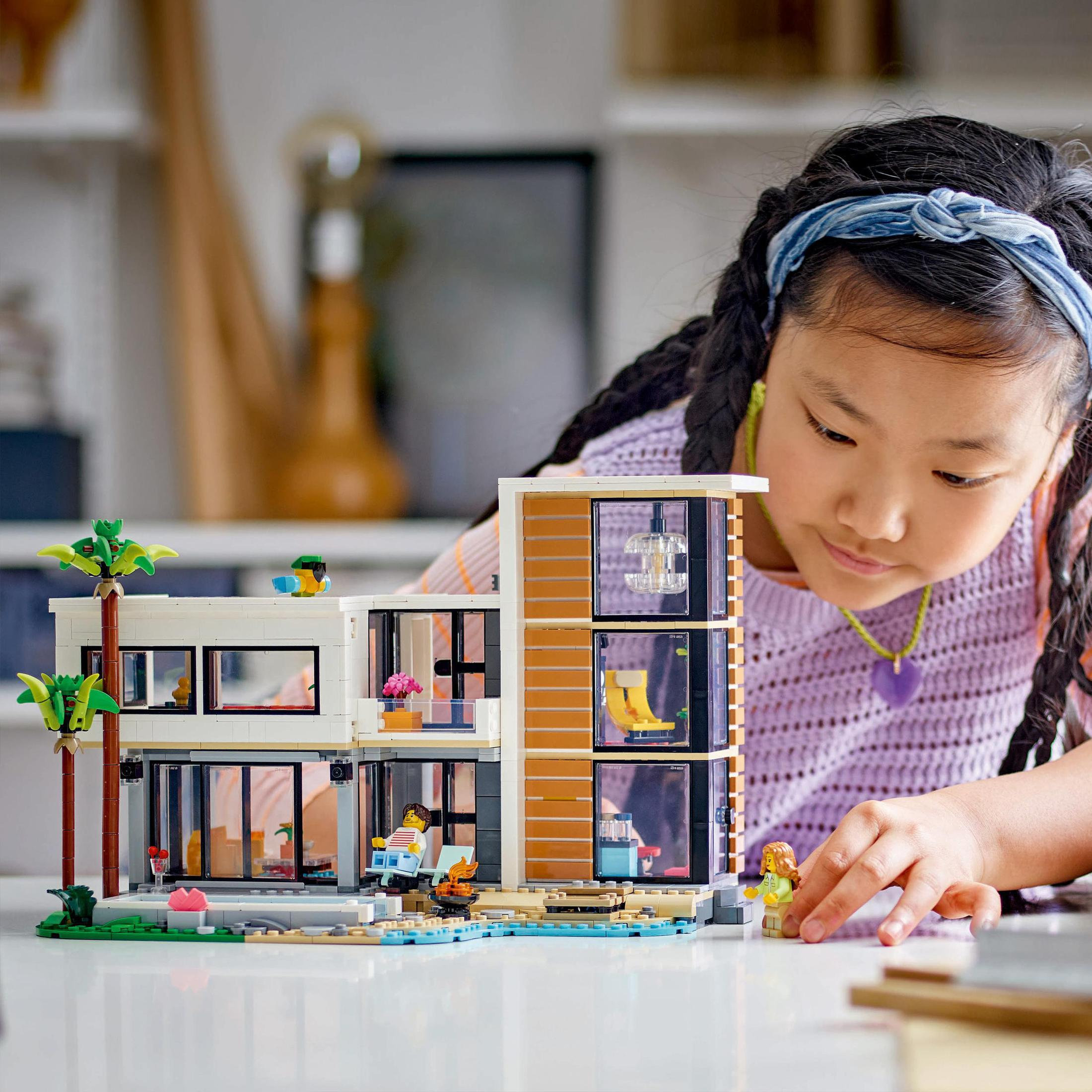 Bir kız Lego evine bakıyor. Bina beyaz, kahverengi ve turuncu. Önünde palmiye ağaçları.
