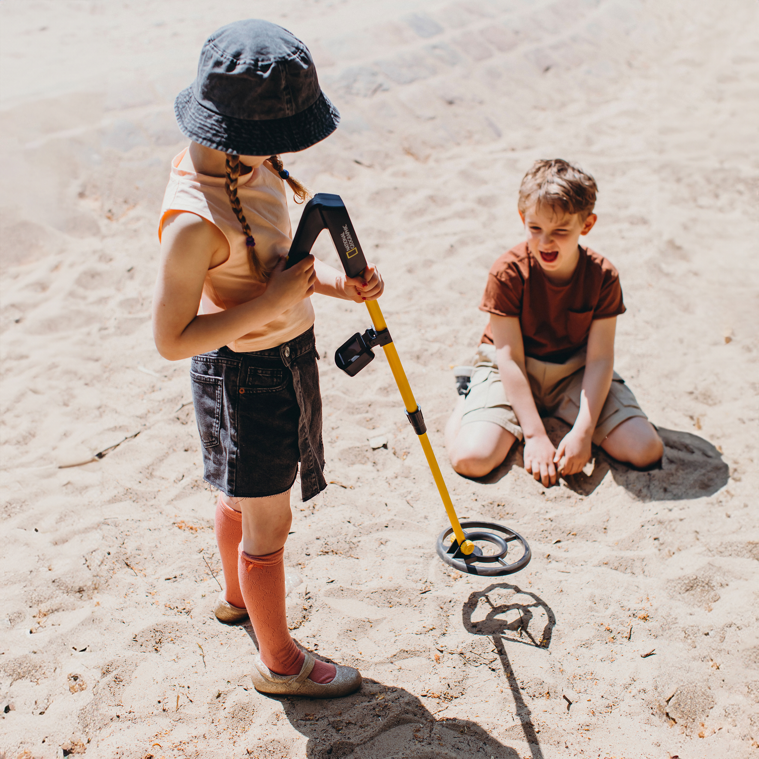 Una niña con sombrero y detector de metales amarillo está de pie en la arena mientras un niño observa.