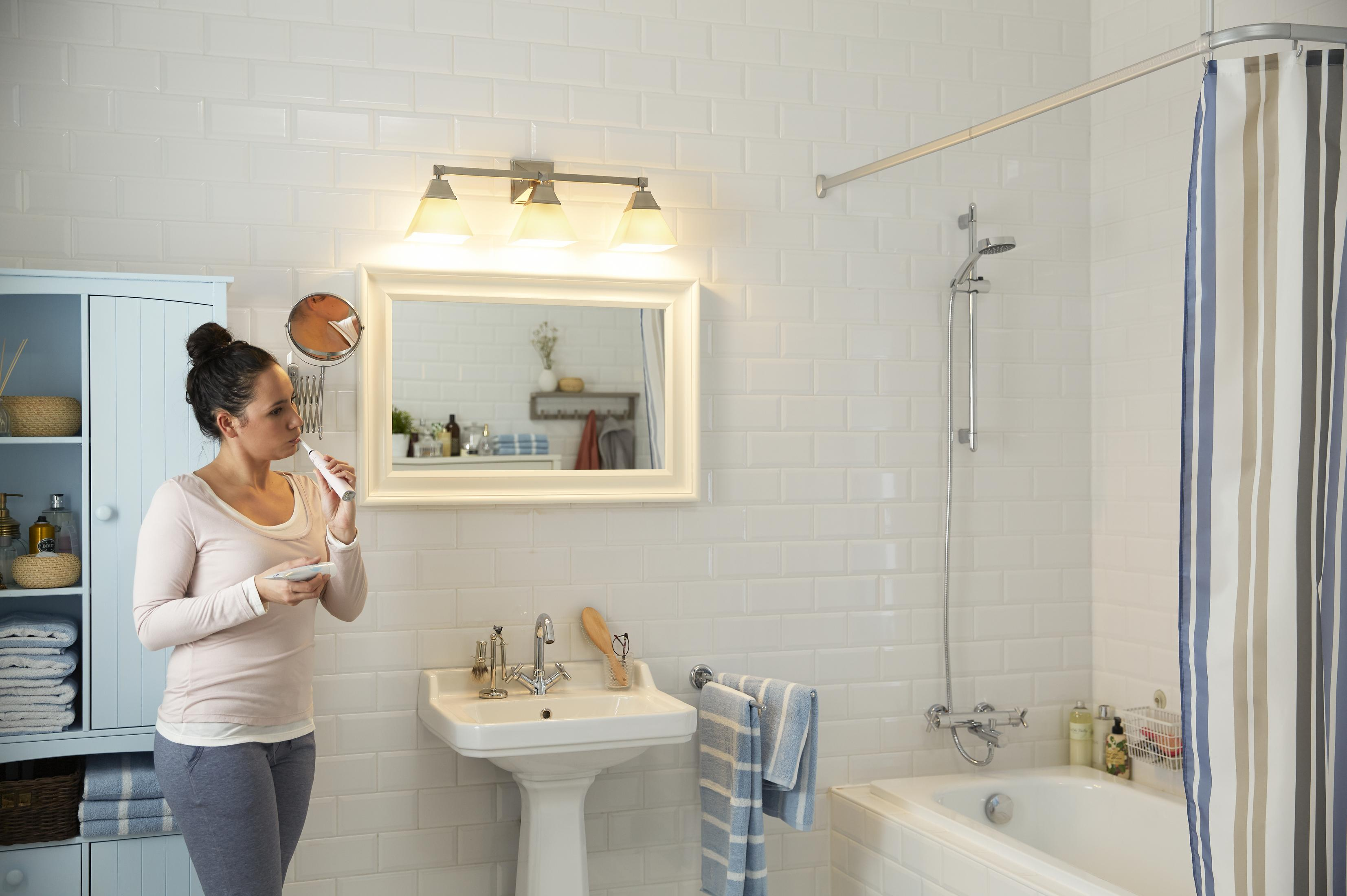 Eine Frau putzt sich in einem weißen Badezimmer die Zähne. Es gibt einen Spiegel, ein Waschbecken und eine Dusche.