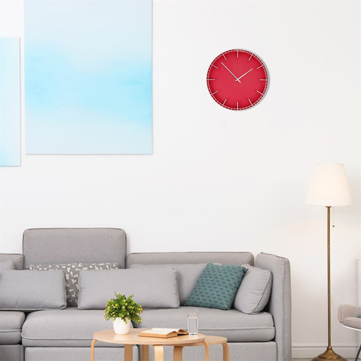 Rote, runde Wanduhr mit weißen Zeigern. Die Uhr hängt an einer weißen Wand über einem grauen Sofa und einer Lampe.