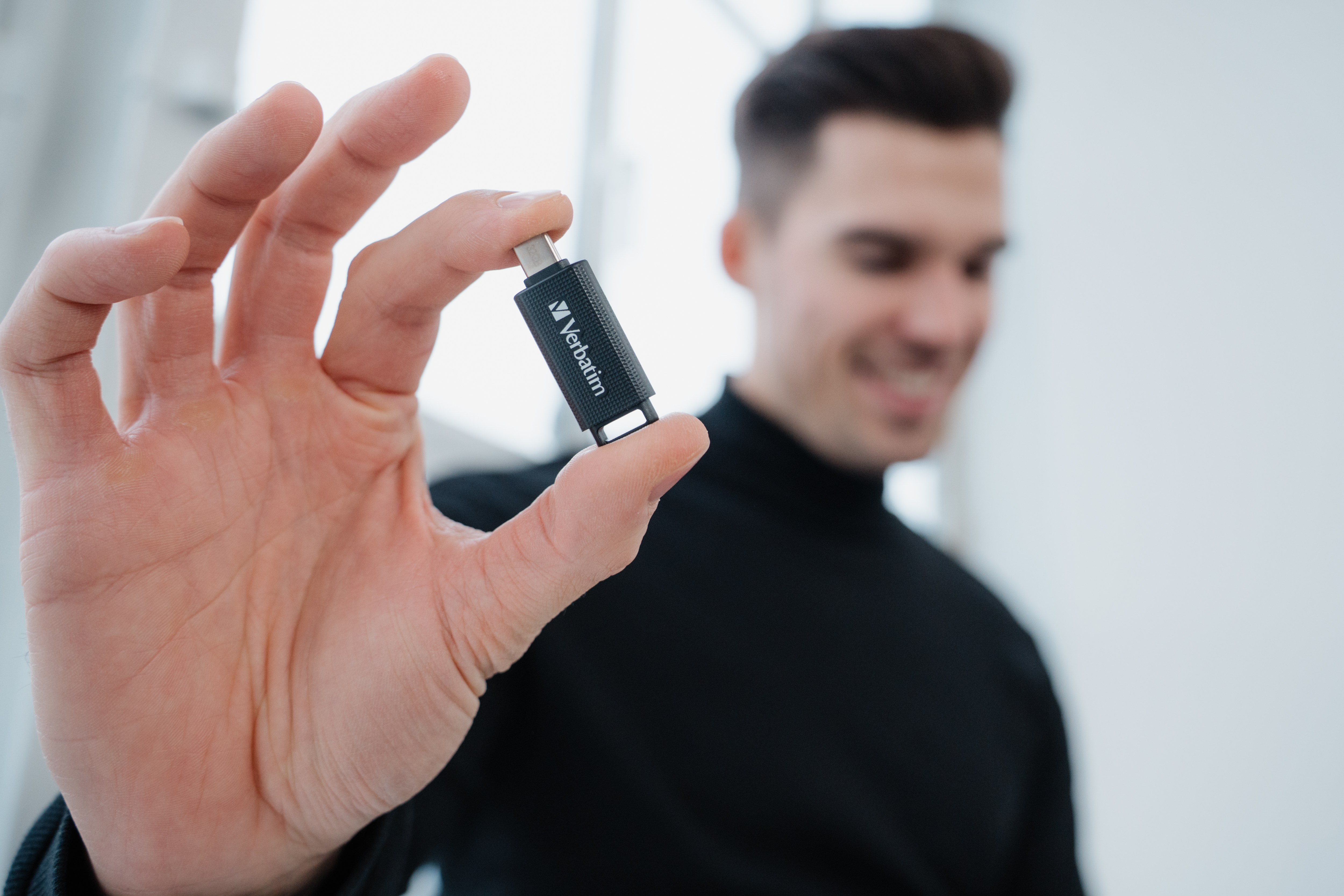 Mężczyzna trzyma czarny pendrive Verbatim. Mężczyzna się uśmiecha. Tło jest białe.
