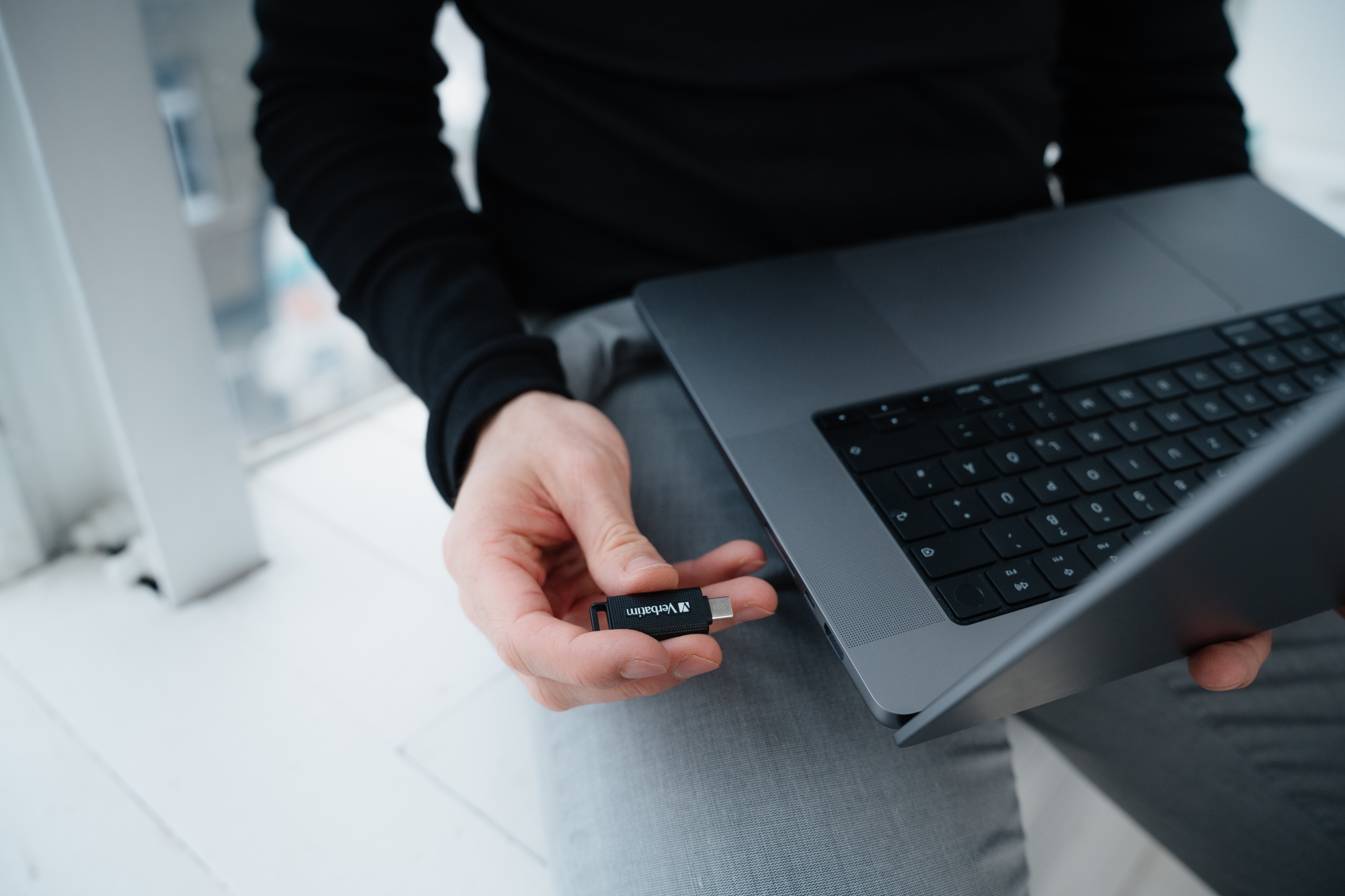 Osoba trzyma czarny pendrive w pobliżu laptopa. Laptop jest otwarty na białej powierzchni.