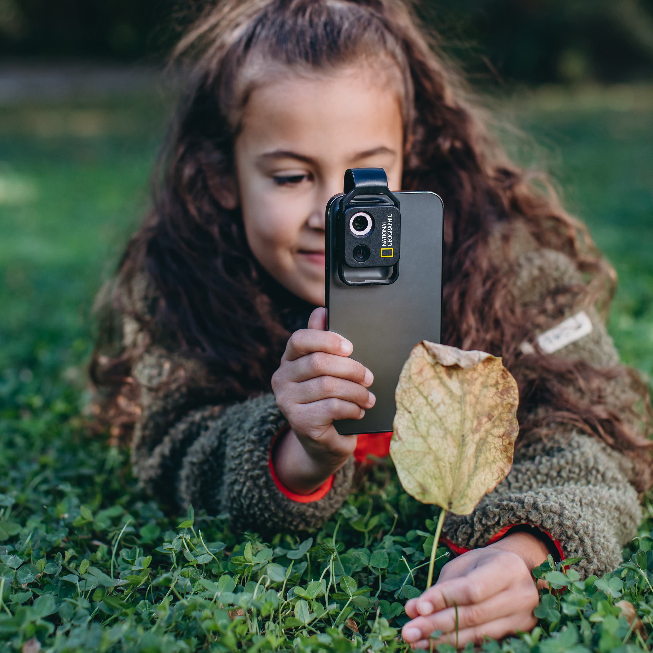 Niña acostada en la hierba, sosteniendo un teléfono con accesorio de cámara, mirando una hoja amarilla.
