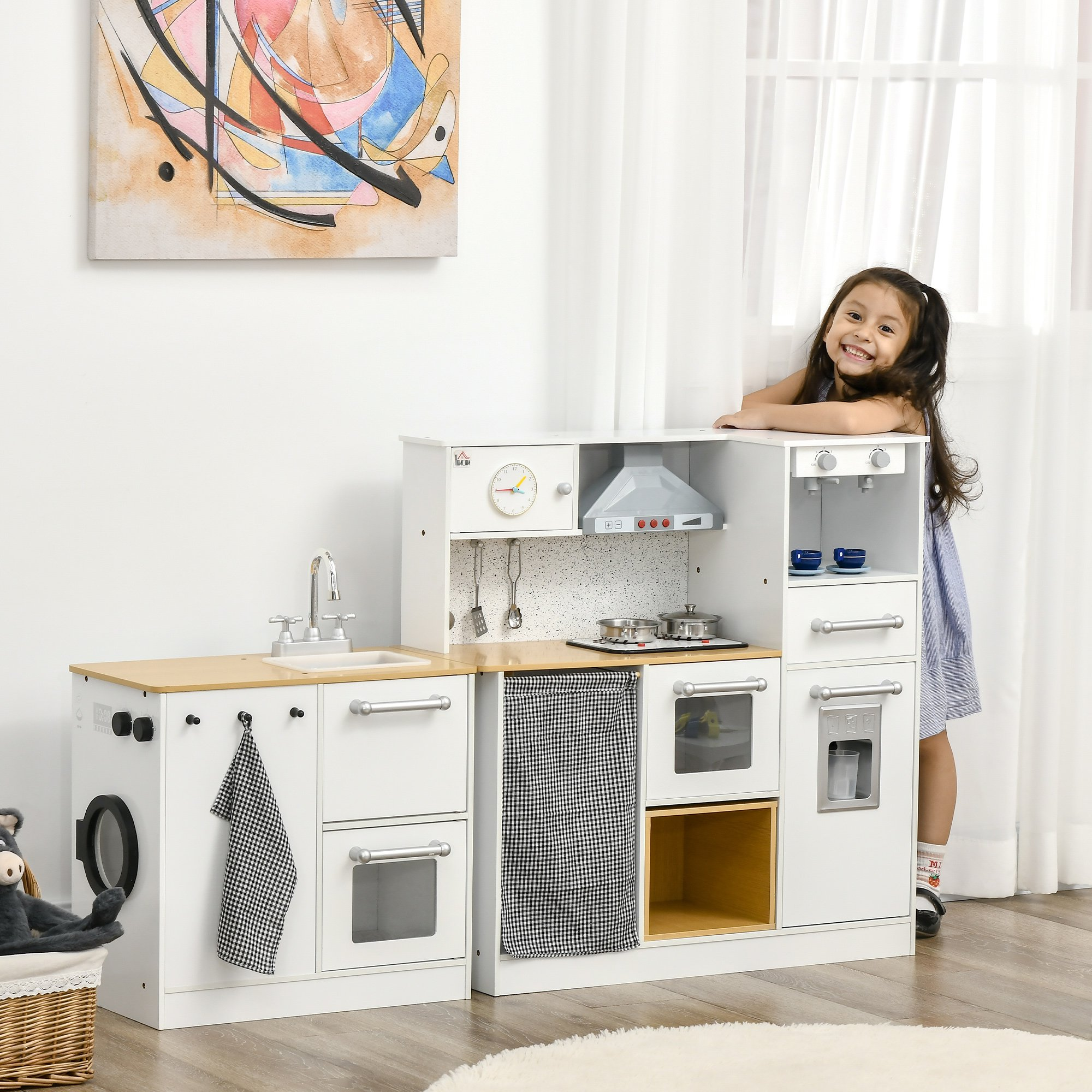 Una niña sonríe junto a una cocina de juguete blanca. Tiene un fregadero, horno y microondas. Cuelga una pintura encima.
