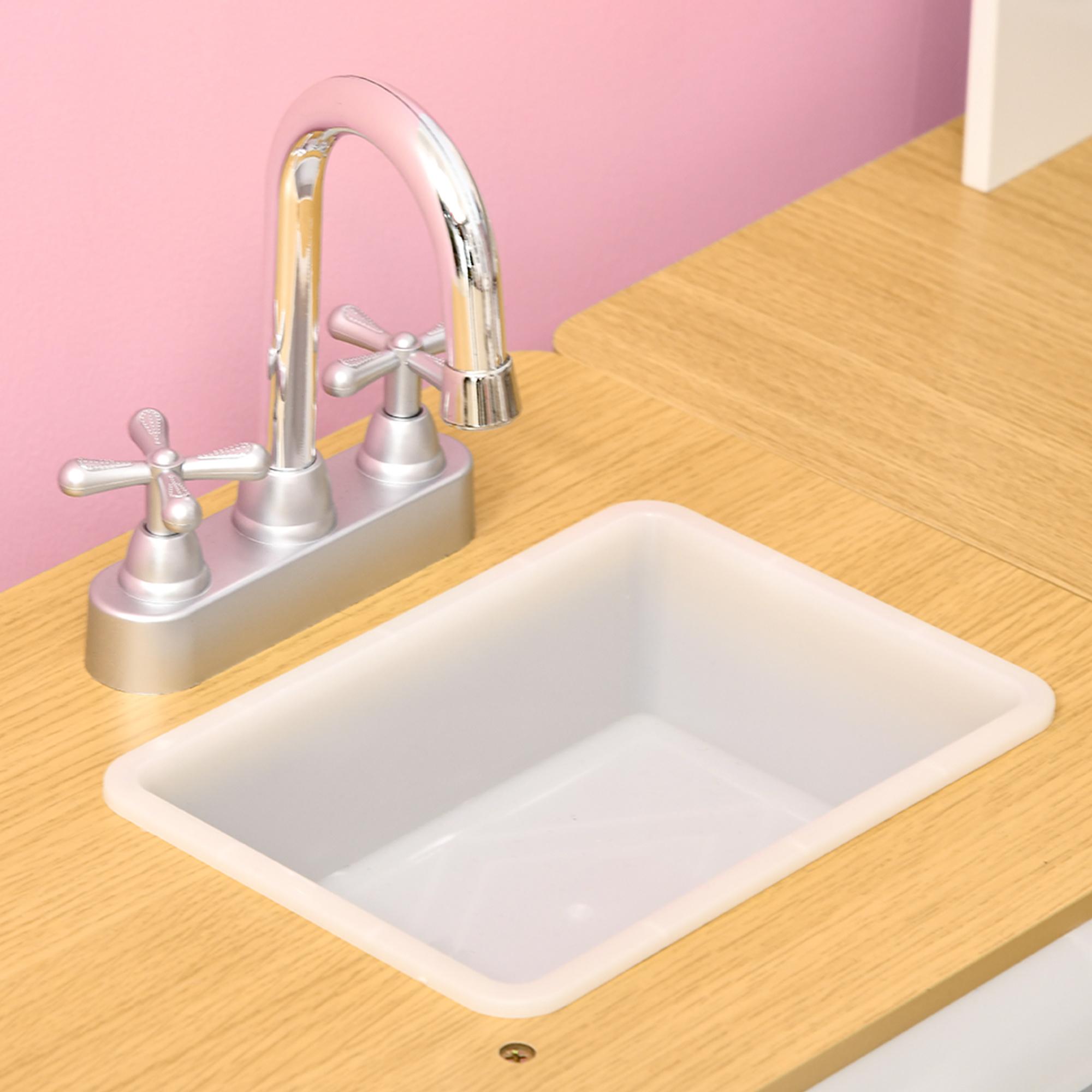 Un fregadero de cocina de juguete con un grifo plateado y un fregadero blanco sobre una encimera de madera, fondo rosa.