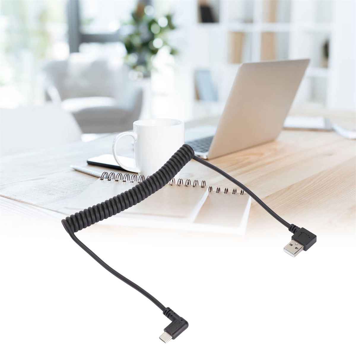 Ein schwarzes USB-C-Kabel mit einem spiralförmigen Abschnitt. Es liegt auf einem Holztisch neben einem Laptop und einem Stift.