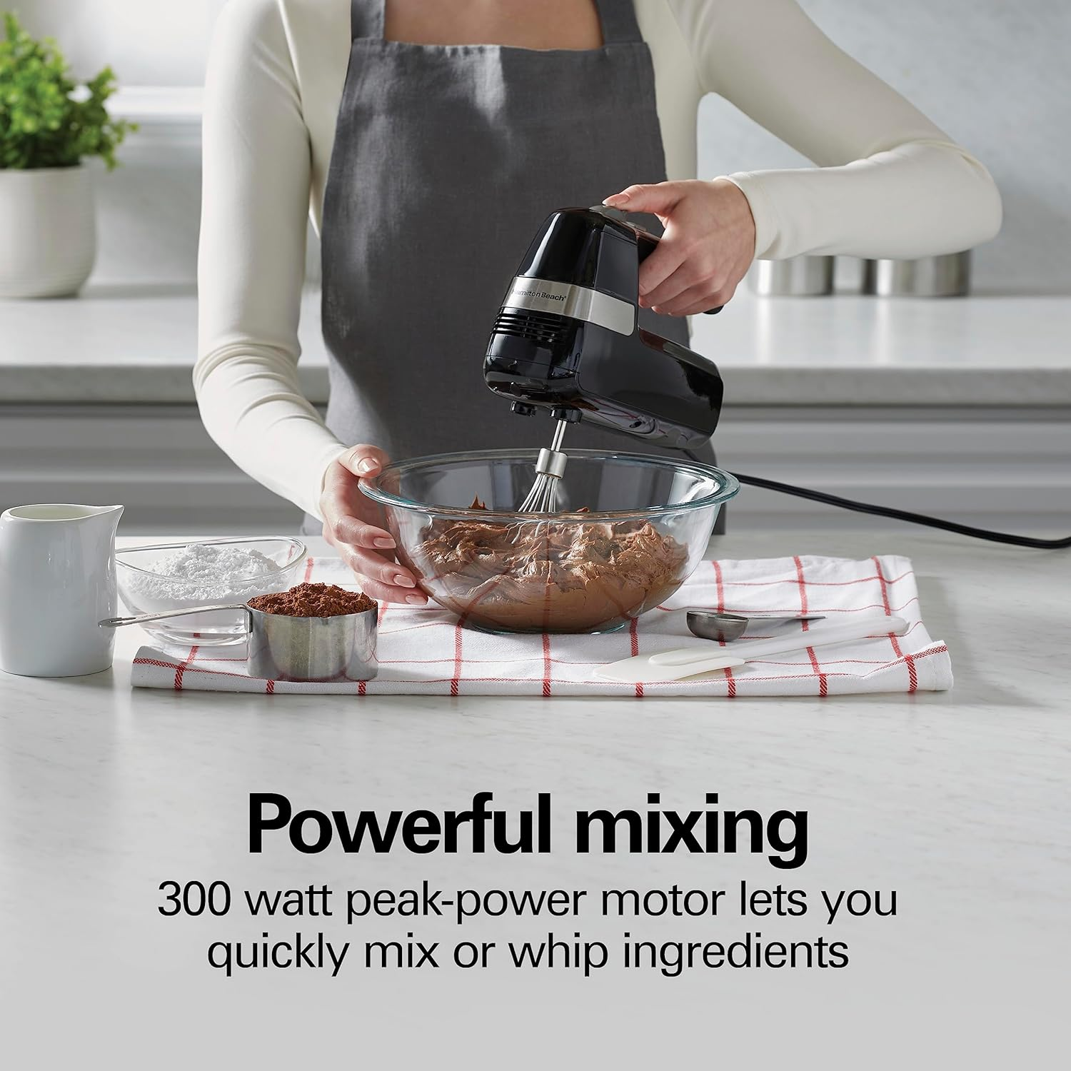 Eine Person, die einen schwarzen Handmixer in einer Glasschüssel verwendet. Zutaten sind sichtbar.
