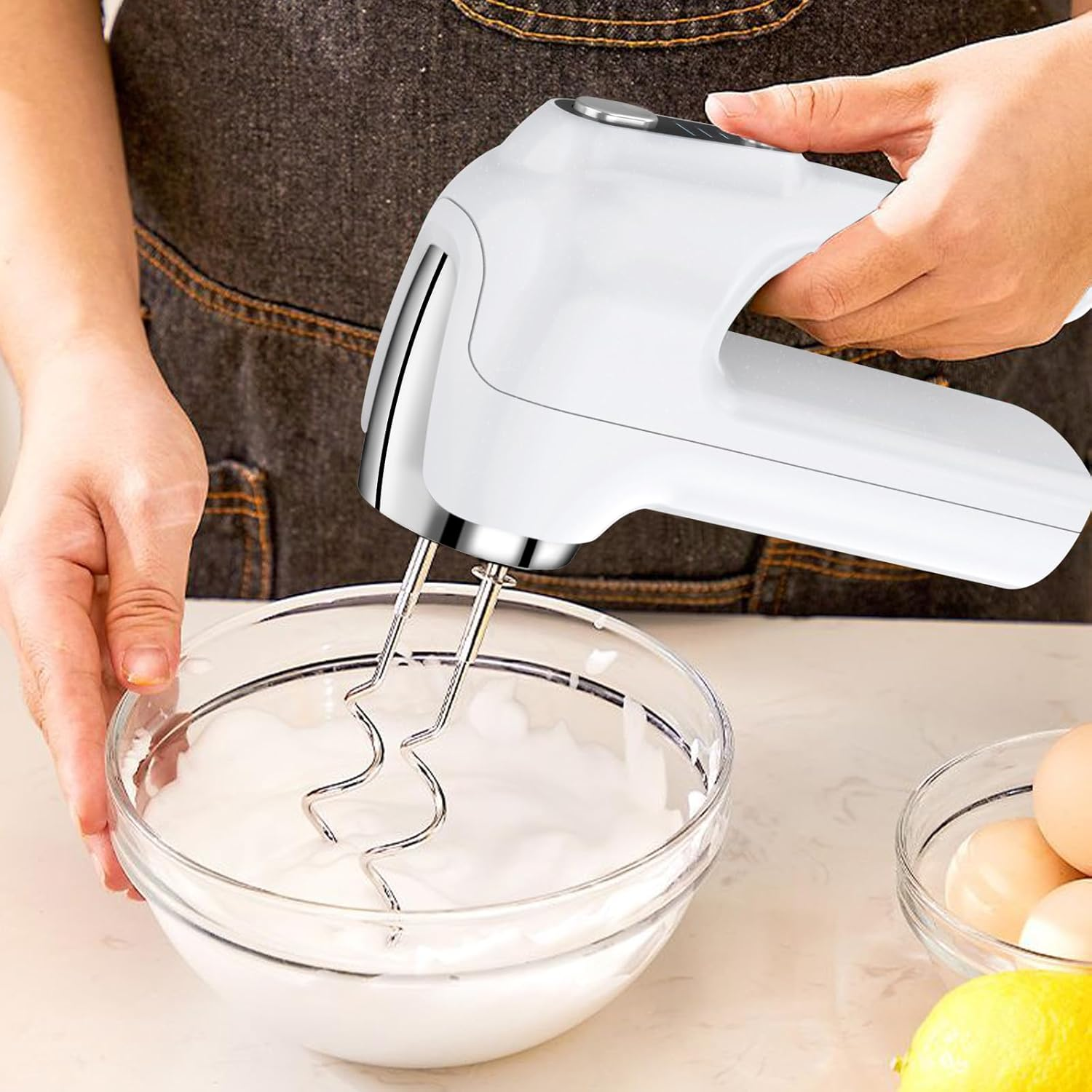 Eine Frau verwendet einen weißen Handmixer, um Sahne in einer Glasschüssel zu schlagen. Eier und eine Zitrone sind in der Nähe.