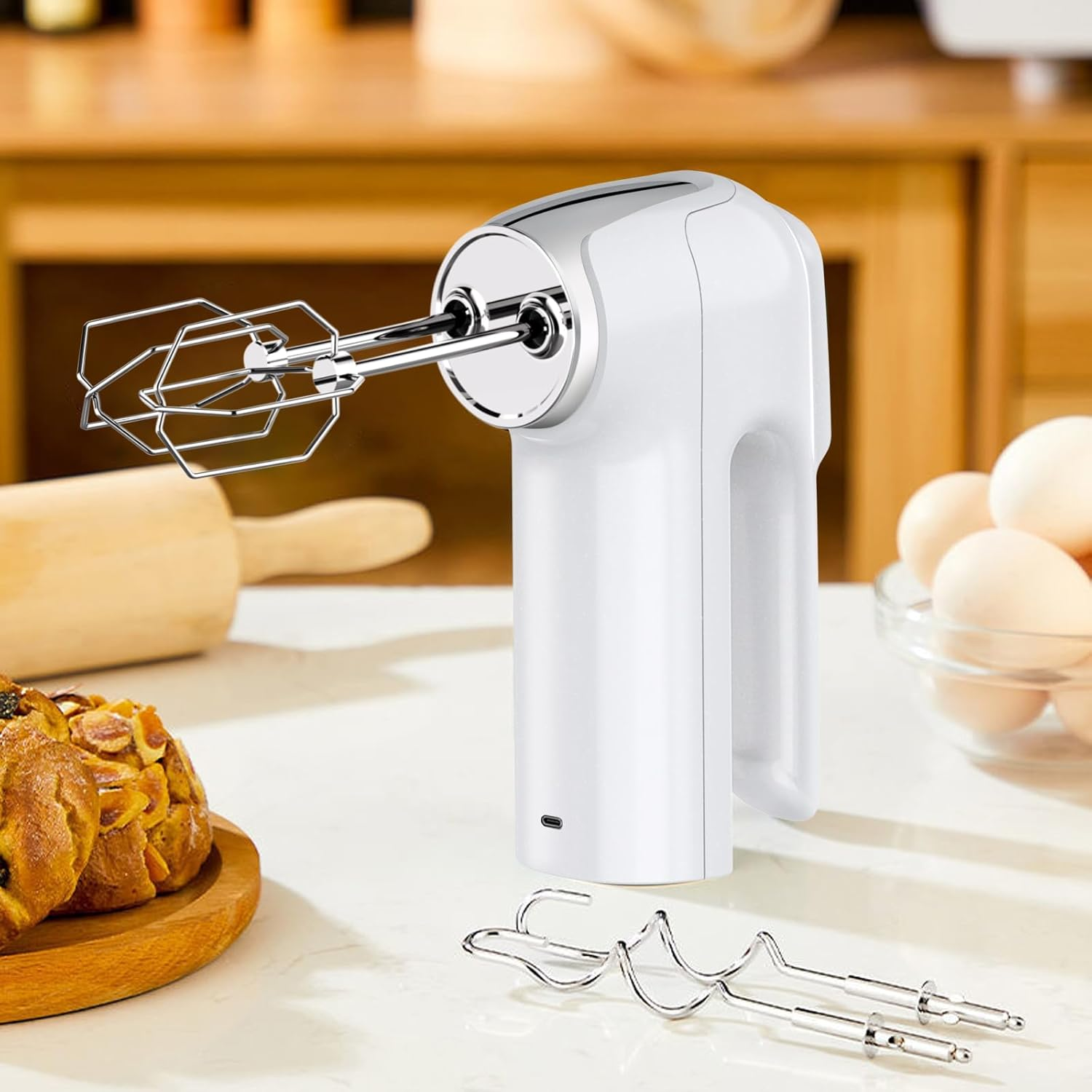 Weißer Handmixer mit Rührbesen auf einer Küchentheke. Brötchen und Eier im Hintergrund.
