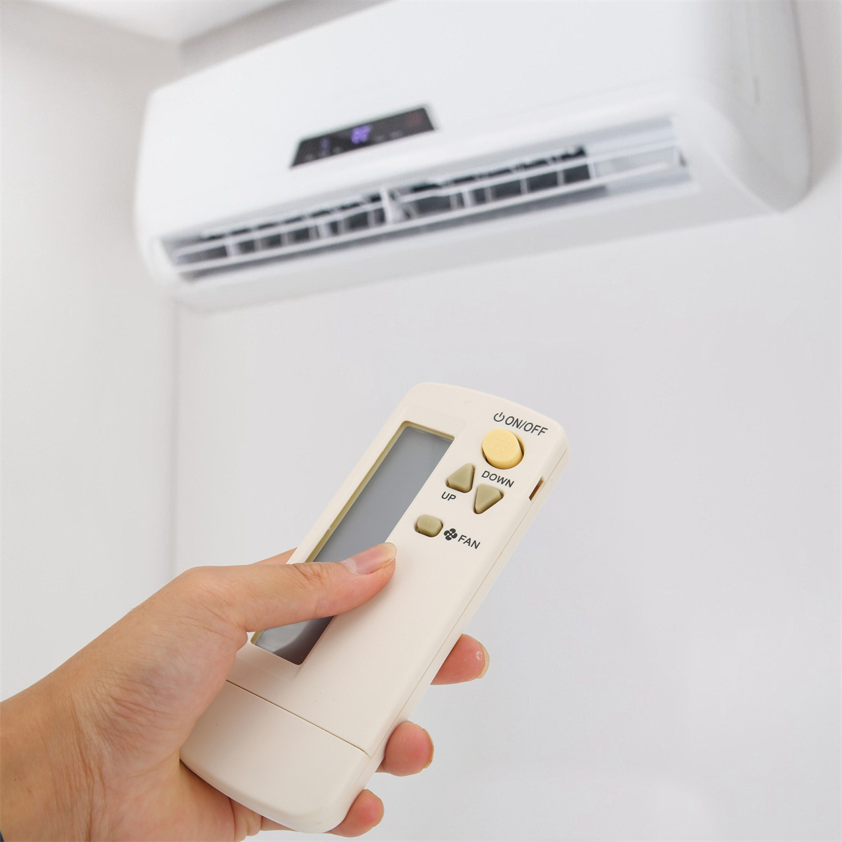 Die Hand einer Person hält eine weiße Fernbedienung. Eine Klimaanlage ist an einer weißen Wand montiert.