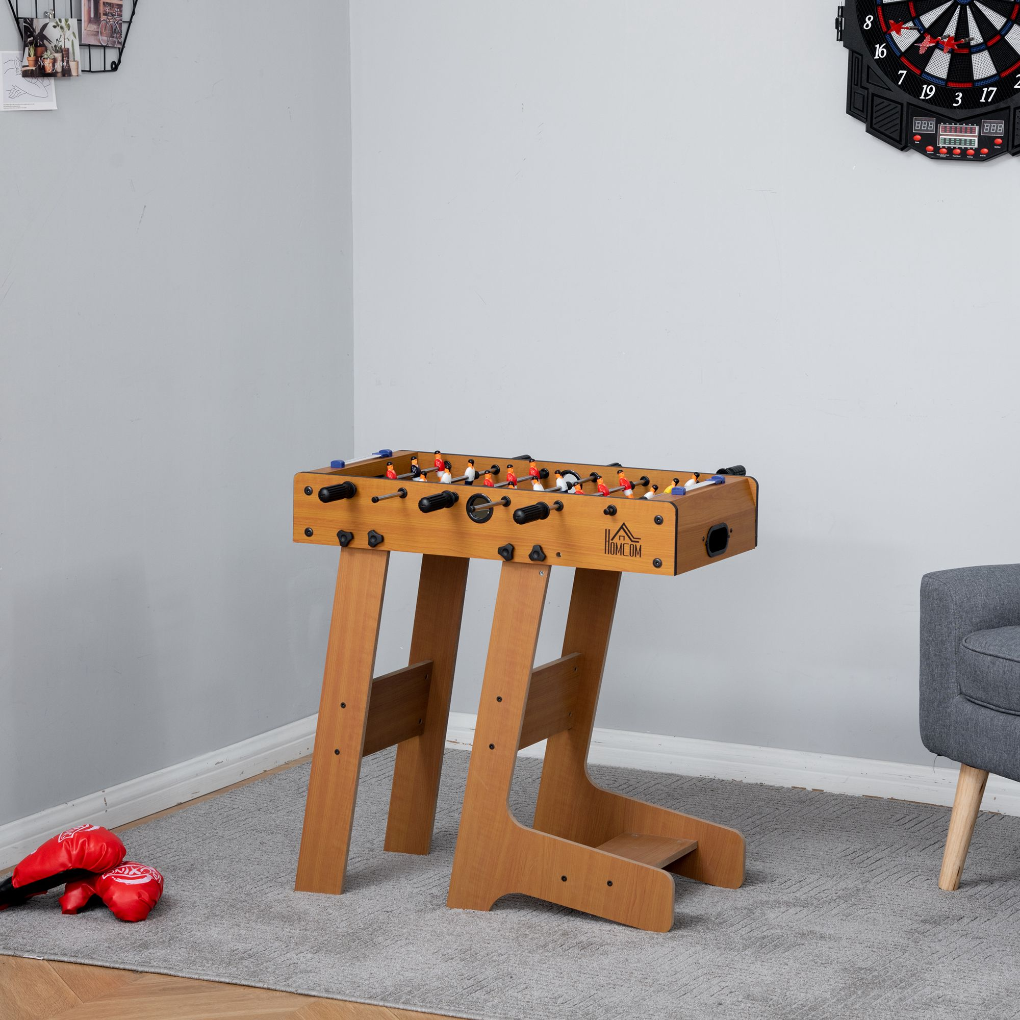Mesa de futbolín sobre un soporte, en una habitación. Alfombra gris, guantes de boxeo rojos, diana en la pared.