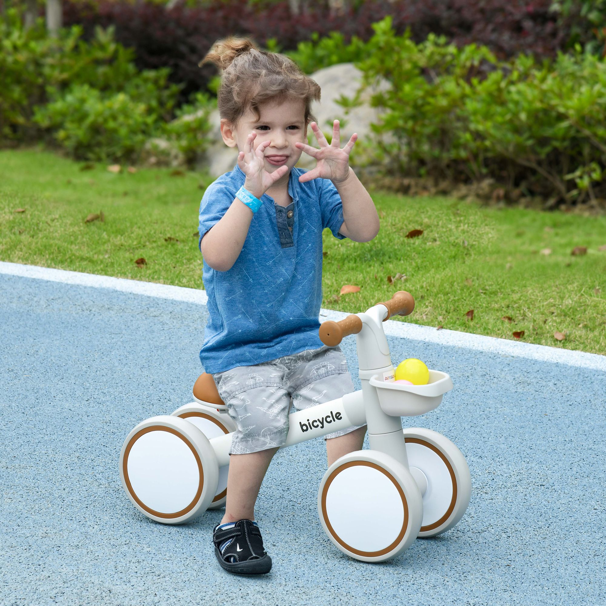 Un niño pequeño en una bicicleta de equilibrio blanca, con una pelota amarilla en la cesta, al aire libre.