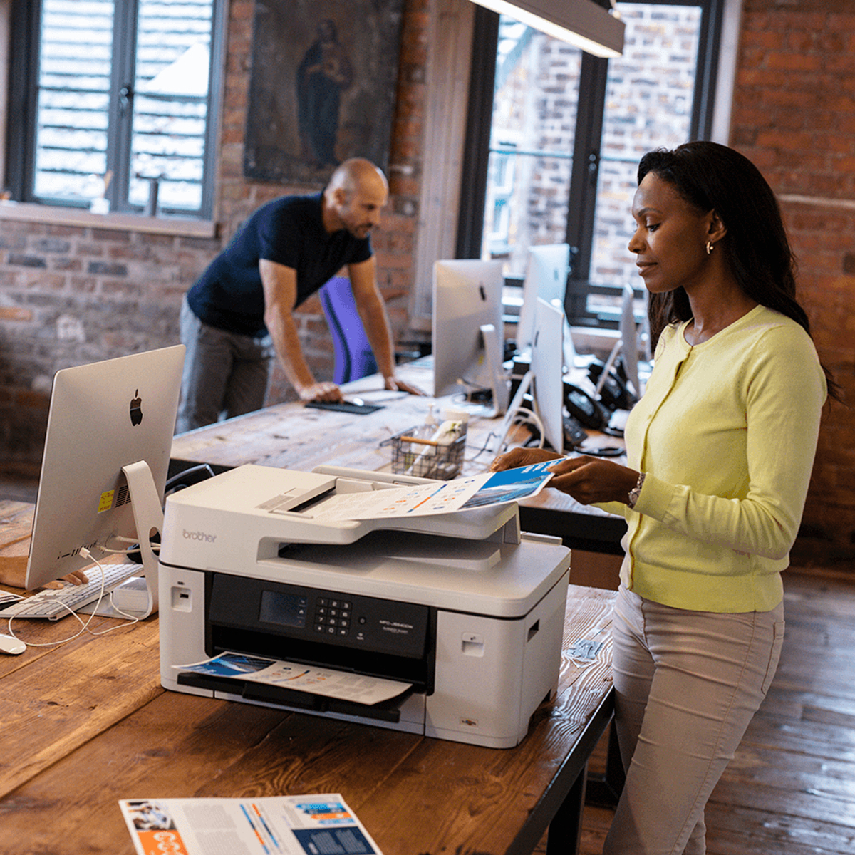 Kobieta i mężczyzna są w biurze. Kobieta używa drukarki, mężczyzna przy biurku.