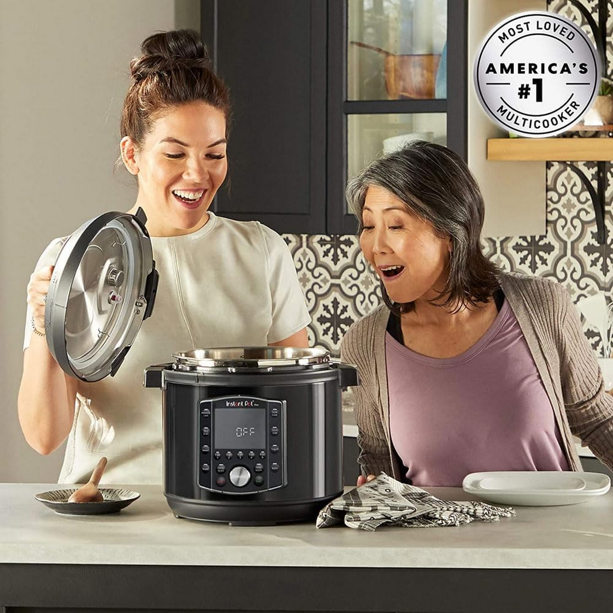 Two women with a black instant pot. One opens the lid. Kitchen background.