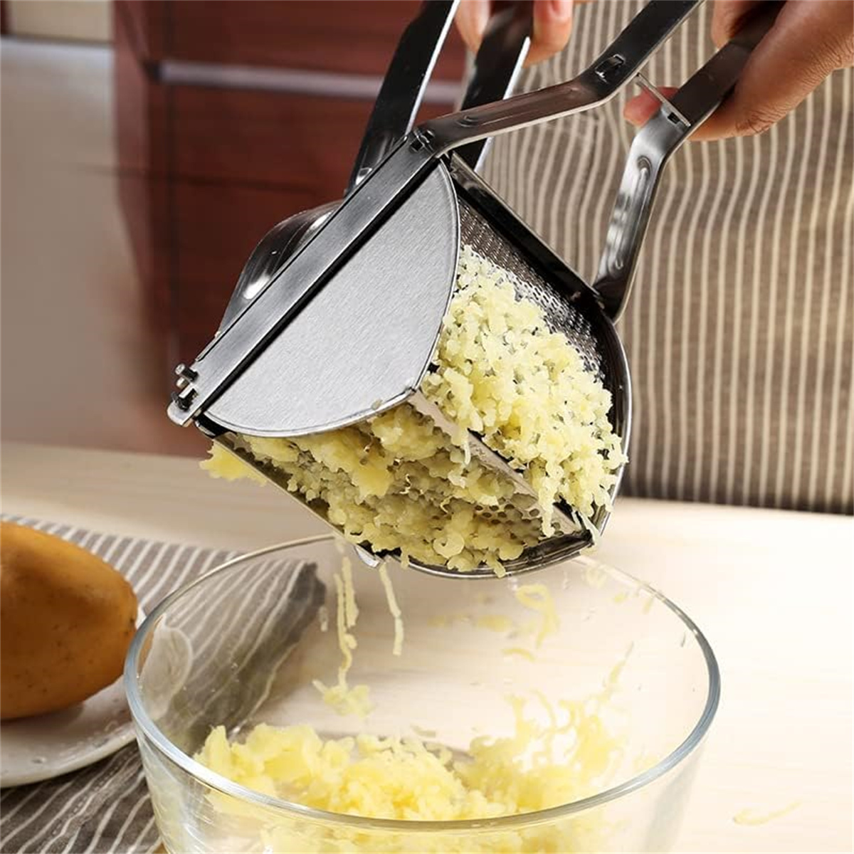 Eine Person presst Kartoffeln durch eine Kartoffelpresse aus Edelstahl in eine Glasschüssel.