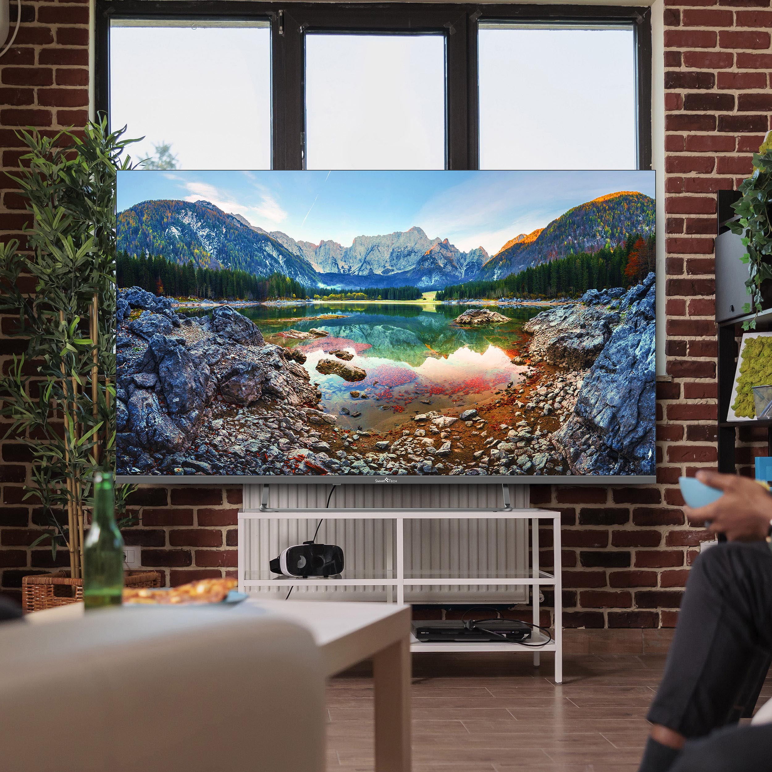 Duży telewizor wyświetlający scenę jeziora górskiego, nad białym stojakiem. Widoczna ceglana ściana i okno.
