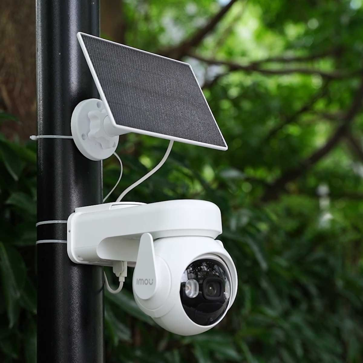 Biała kamera bezpieczeństwa z panelem słonecznym zamontowana na czarnym słupie w zielonym otoczeniu.
