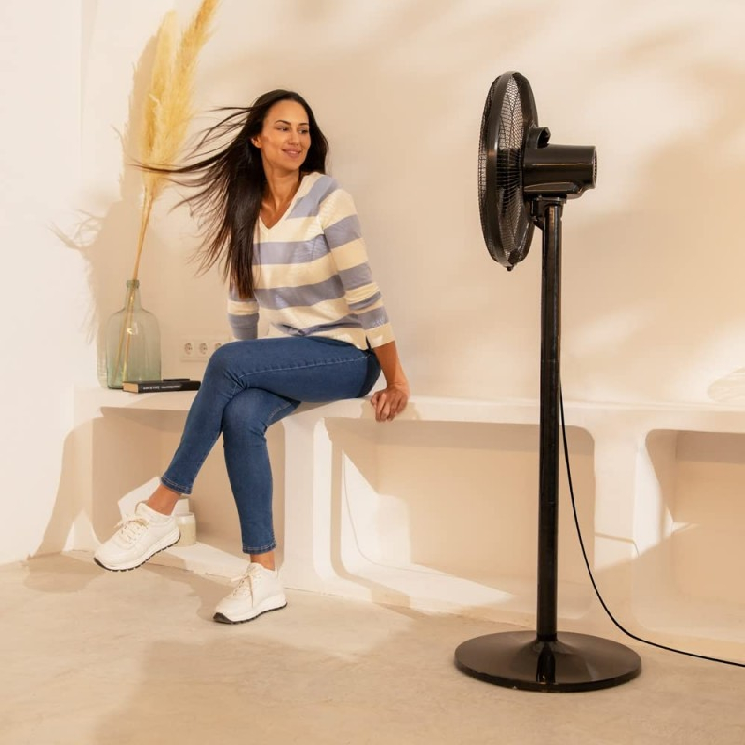 Kobieta siedząca obok czarnego wentylatora. Ma na sobie koszulę w paski i dżinsy, uśmiecha się w jasnym pokoju.