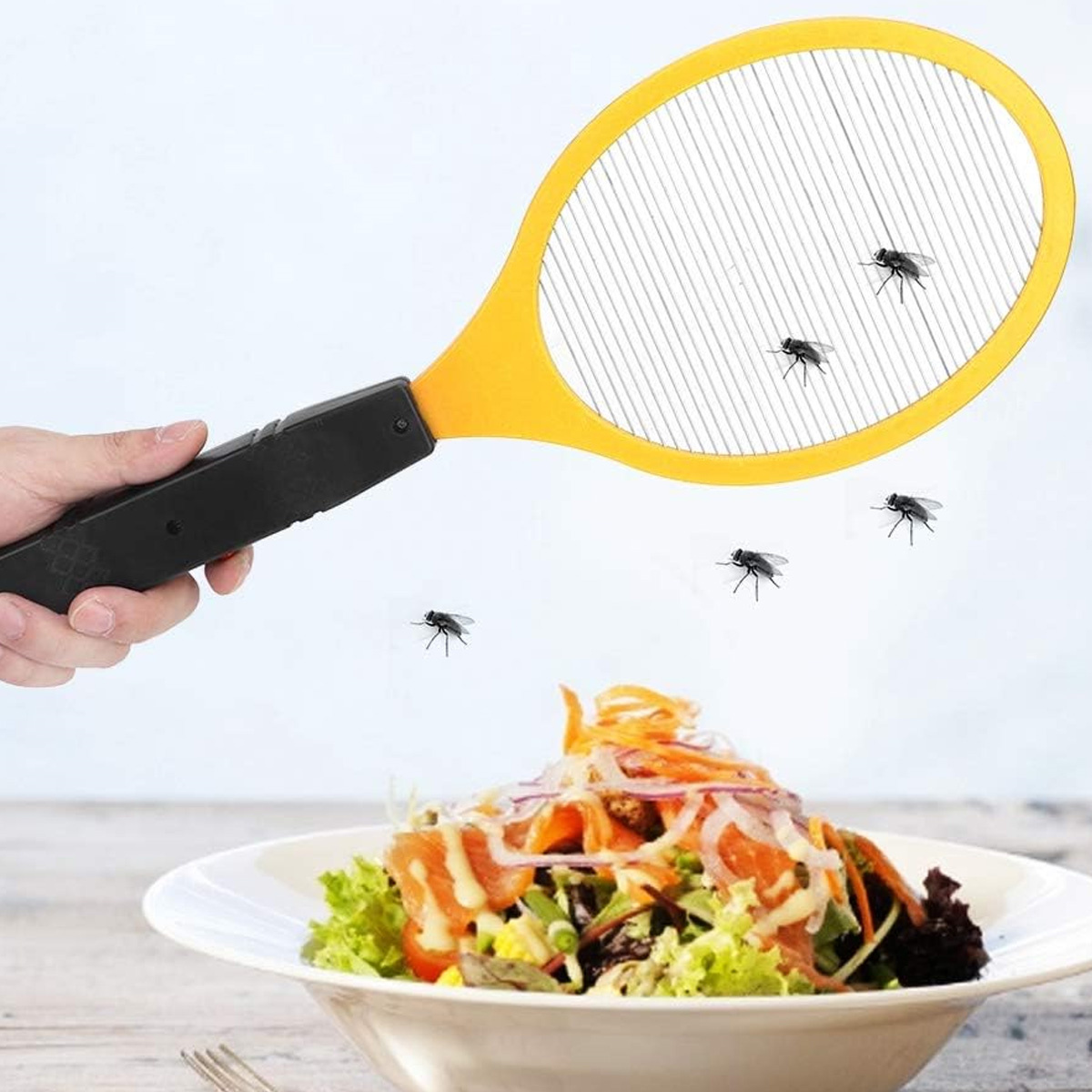 Hand hält einen gelben elektrischen Fliegenklatscher über einem Salat mit Fliegen.