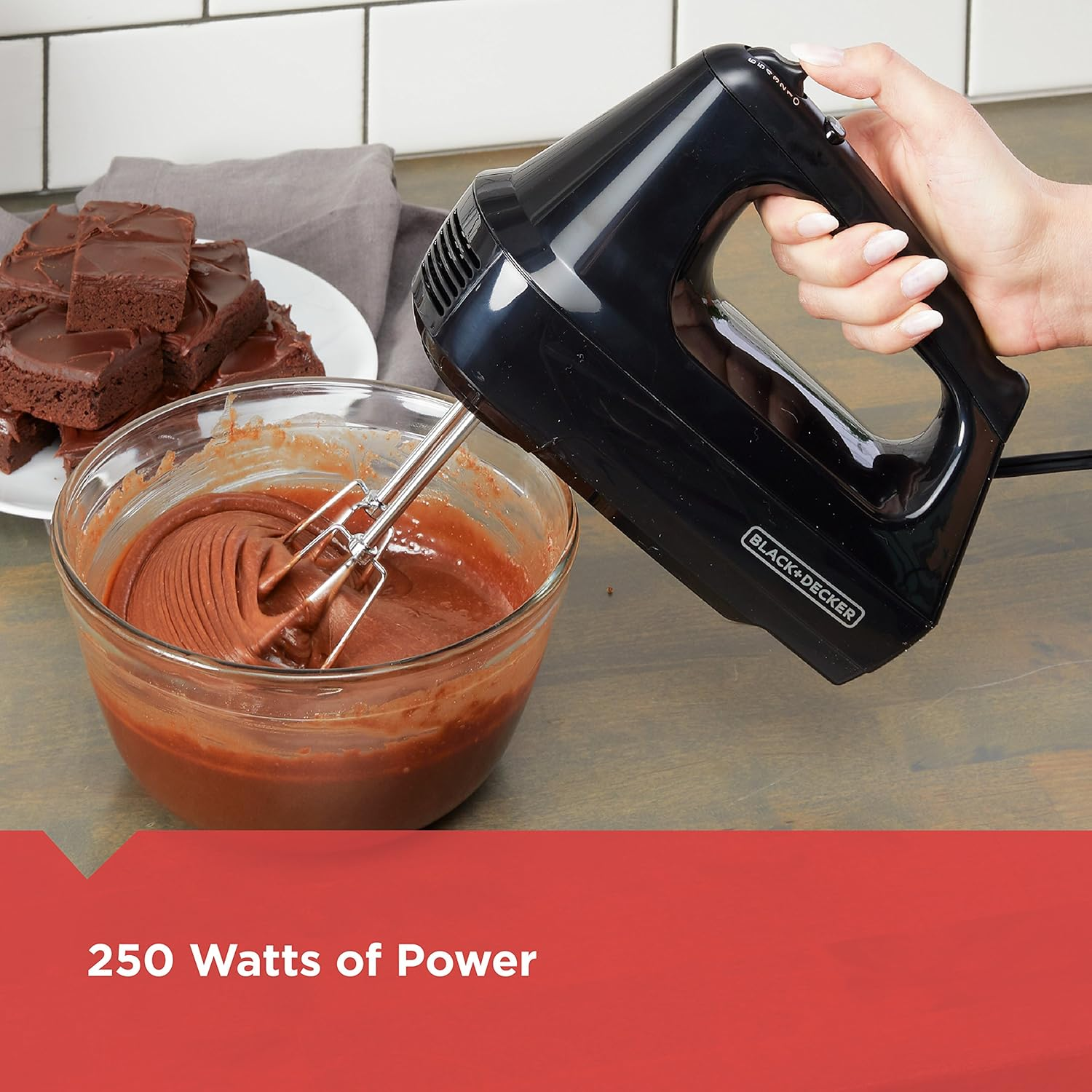 Eine Person verrührt Teig mit einem schwarzen Handmixer, einer Glasschüssel und Brownies im Hintergrund.