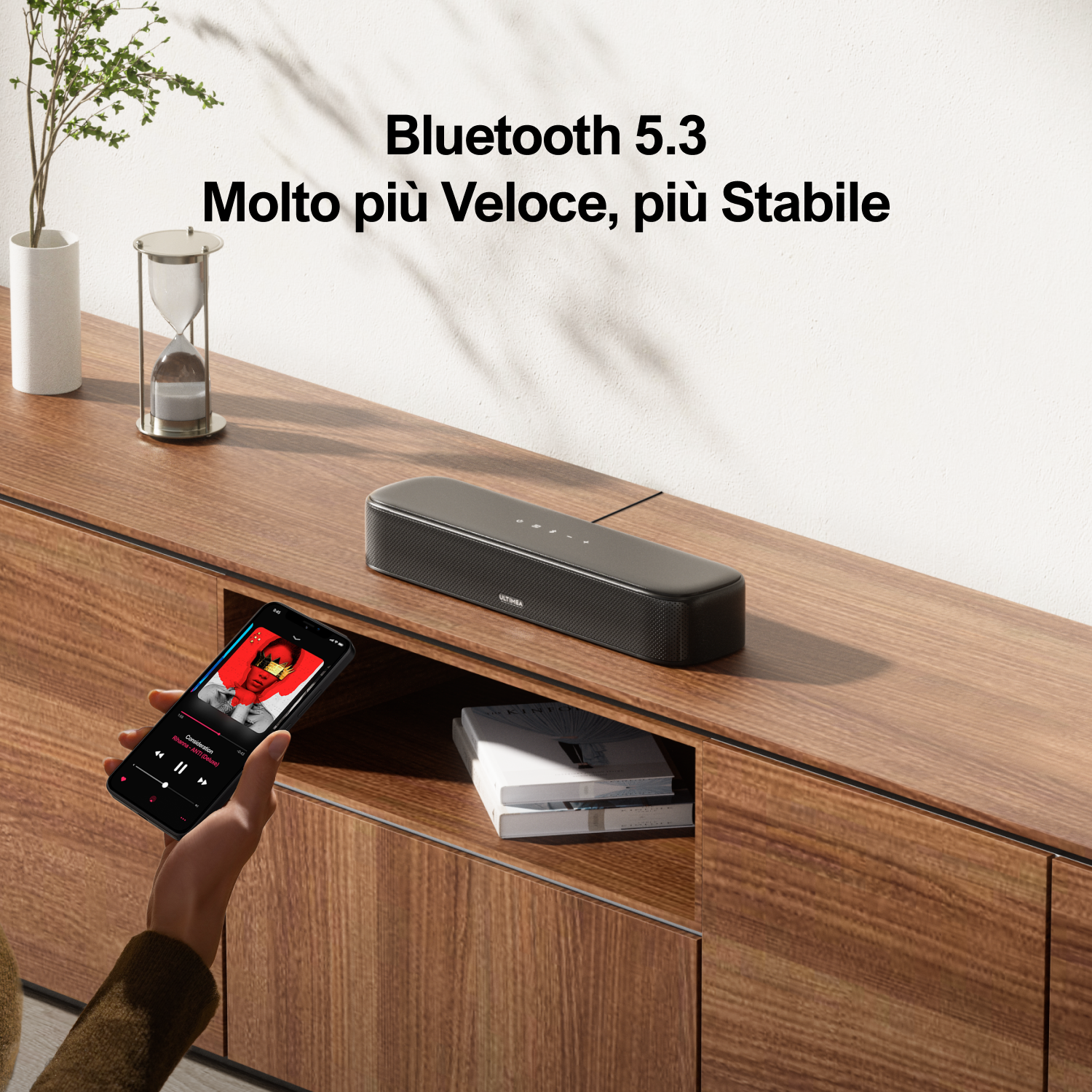 Una soundbar nera è appoggiata su un mobile in legno, con un telefono che mostra i controlli della musica in primo piano.