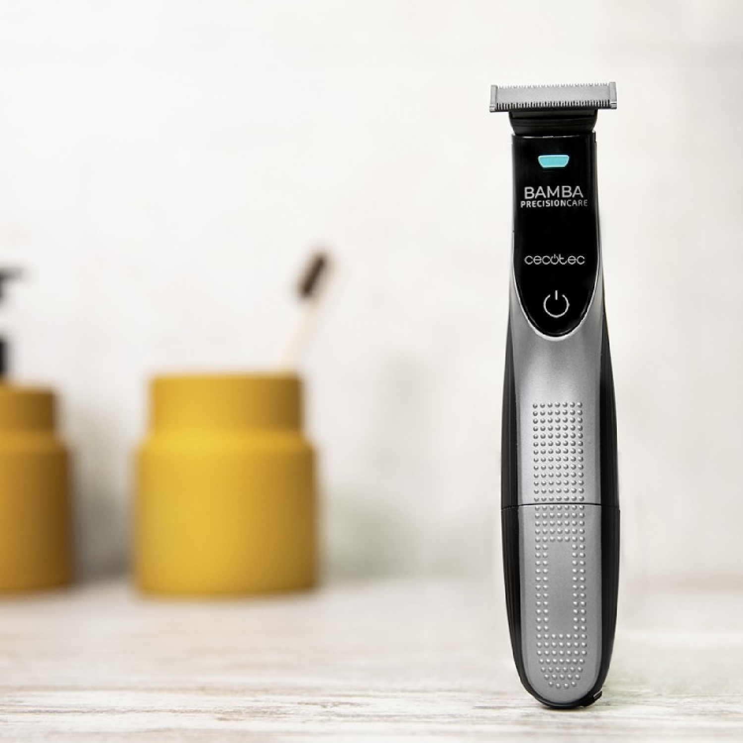 Afeitadora eléctrica negra y plateada. El nombre del producto es Bamba PrecisionCare. Está sobre una superficie de madera cerca de objetos amarillos.