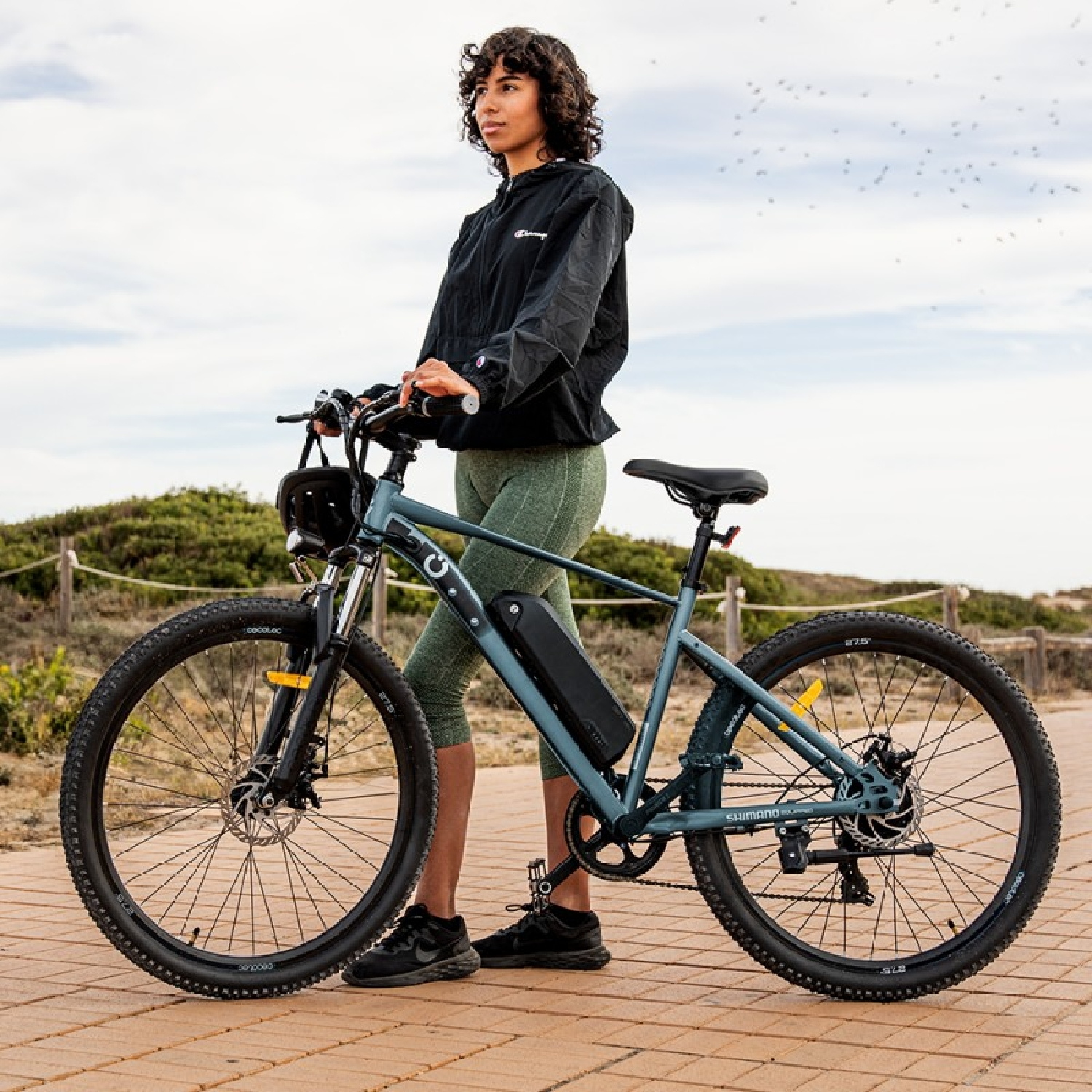 Mujer con bicicleta eléctrica en un paseo marítimo cerca de la playa. La bicicleta es gris, y ella lleva una chaqueta negra y leggings.