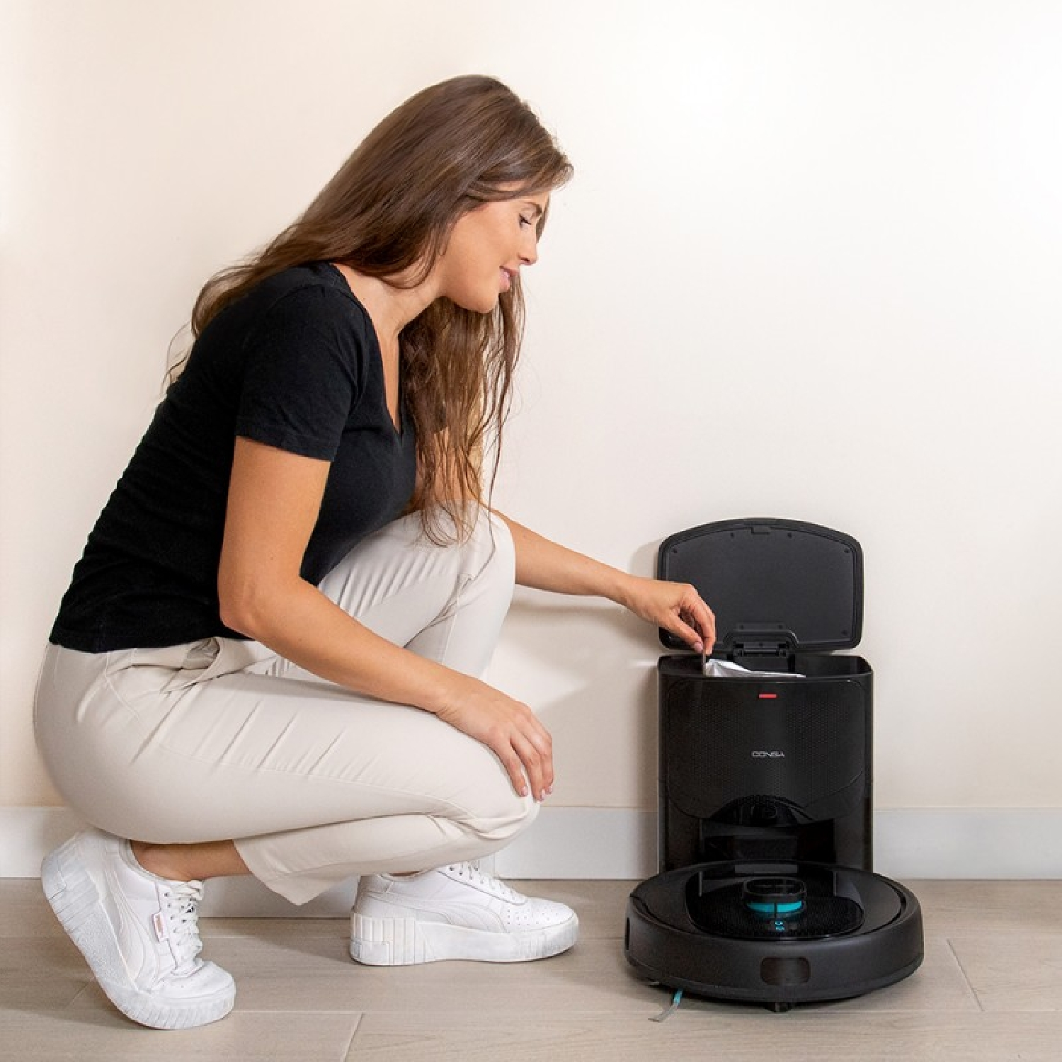 Donna che mette un sacchetto nel contenitore della polvere di un robot aspirapolvere nero. È in una stanza con pareti bianche.