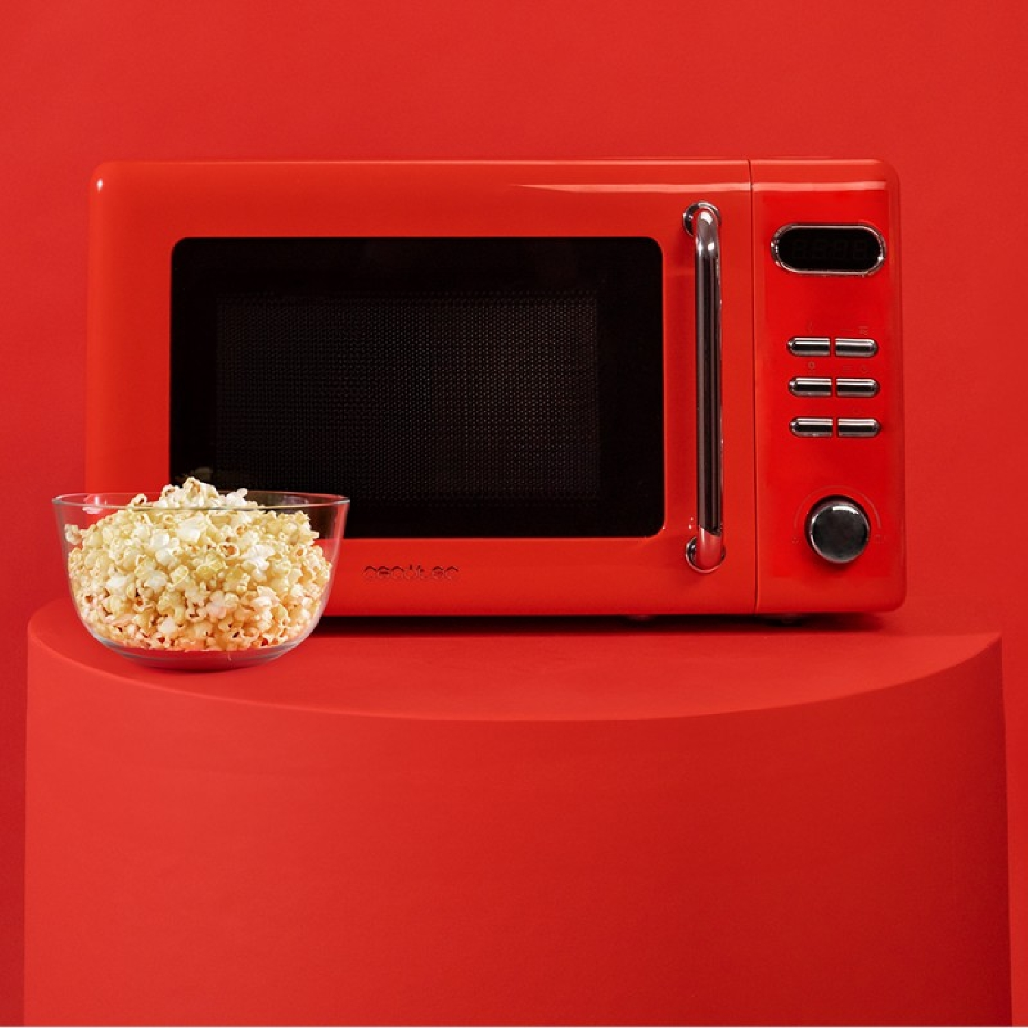 Czerwona kuchenka mikrofalowa z popcornem w szklanej misce, na czerwonym cokole, na czerwonym tle.